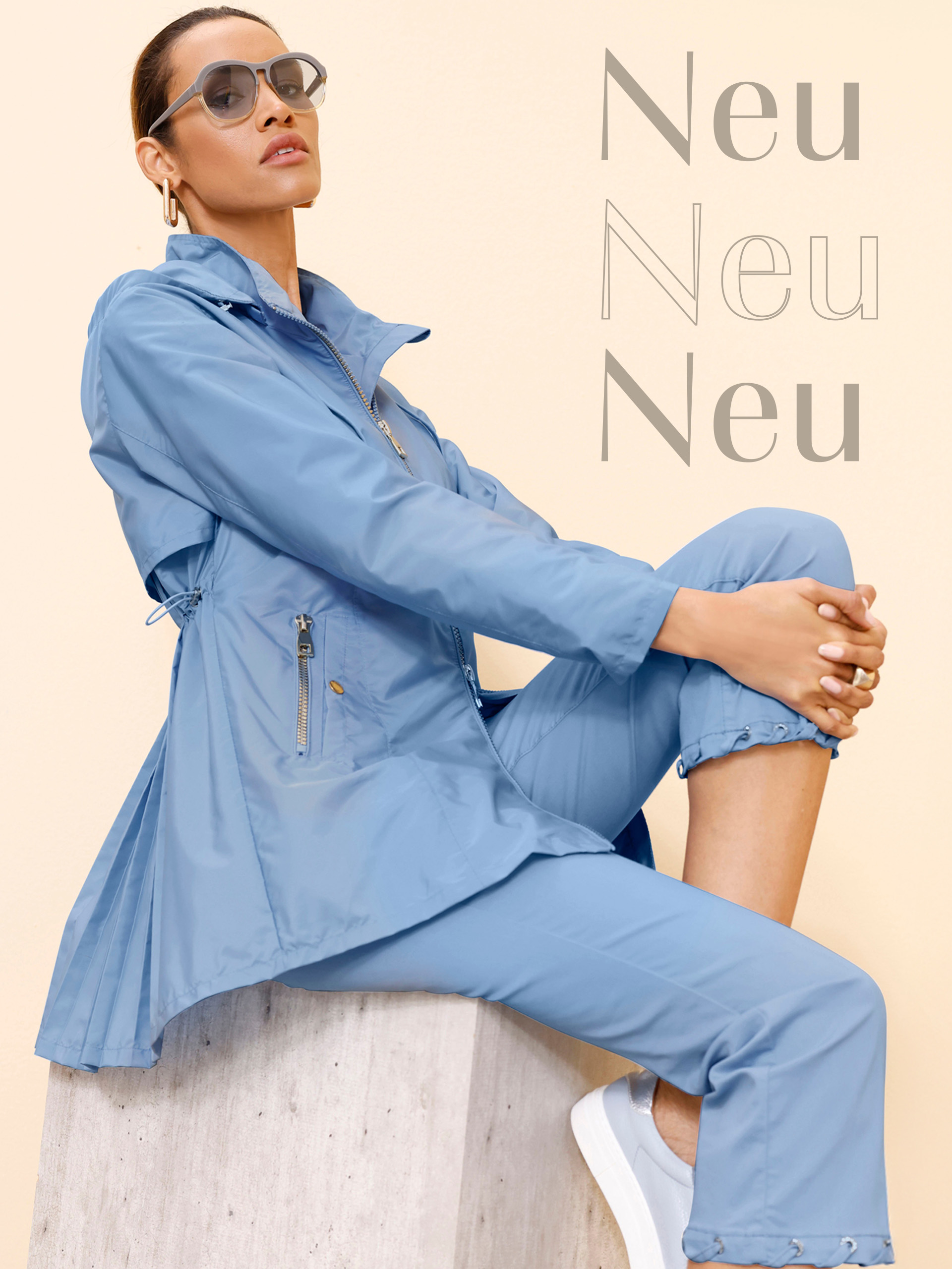 Das Bild zeigt eine Frau, die eine hellblaue Freizeitjacke und passende Hosen trägt. Sie sitzt auf einer hellbeigen Fläche und trägt eine Sonnenbrille. Auf dem Bildes steht dreimal das Wort "Neu" in verschiedenen Grautönen und Schriftstilen.