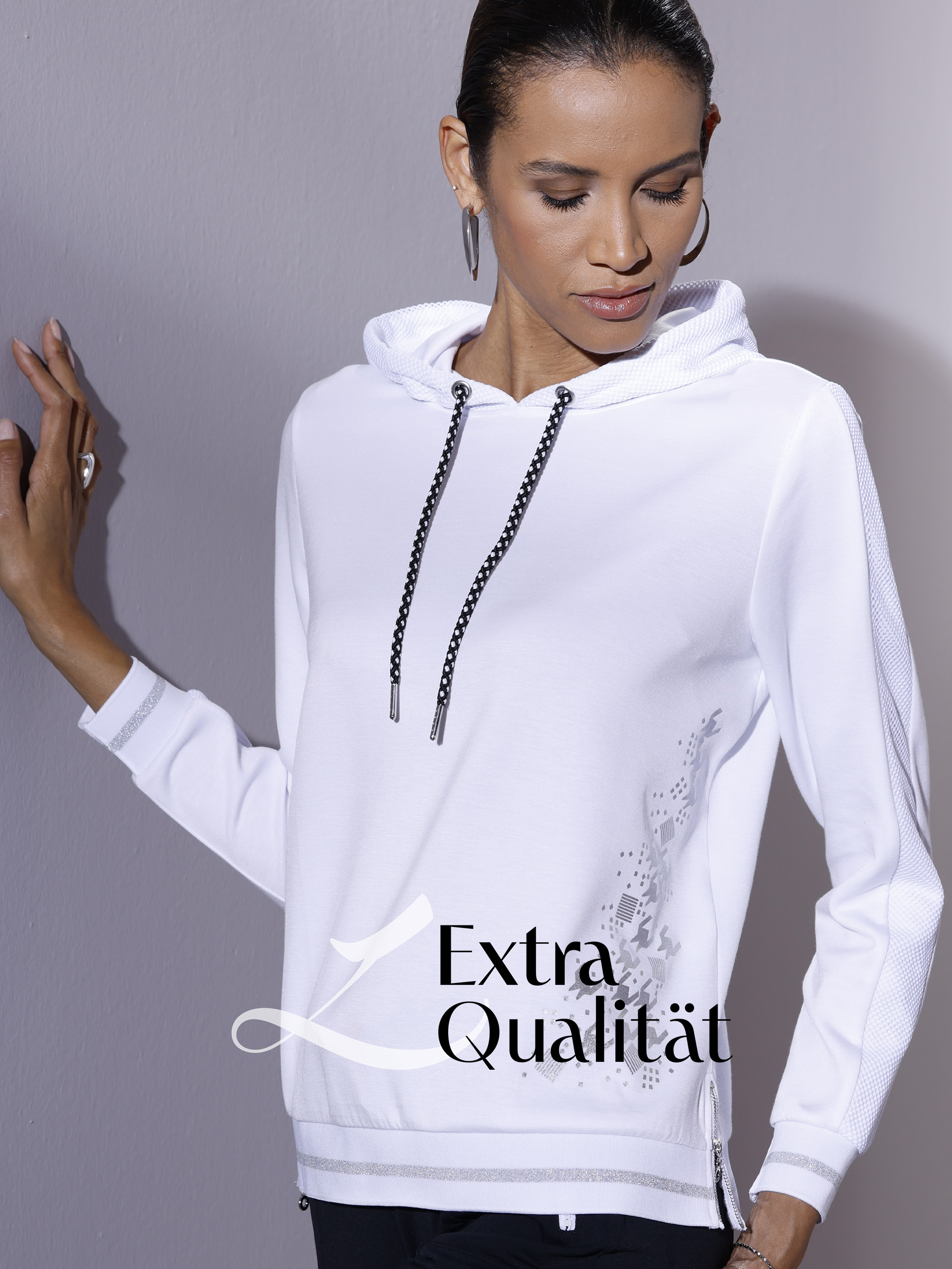 Eine Frau in weißem Kapuzenpullover. Der Pulli hat verzierte Schnüre und Ärmel. "Extra Qualität" und ein stilisiertes "L" sind zu sehen.