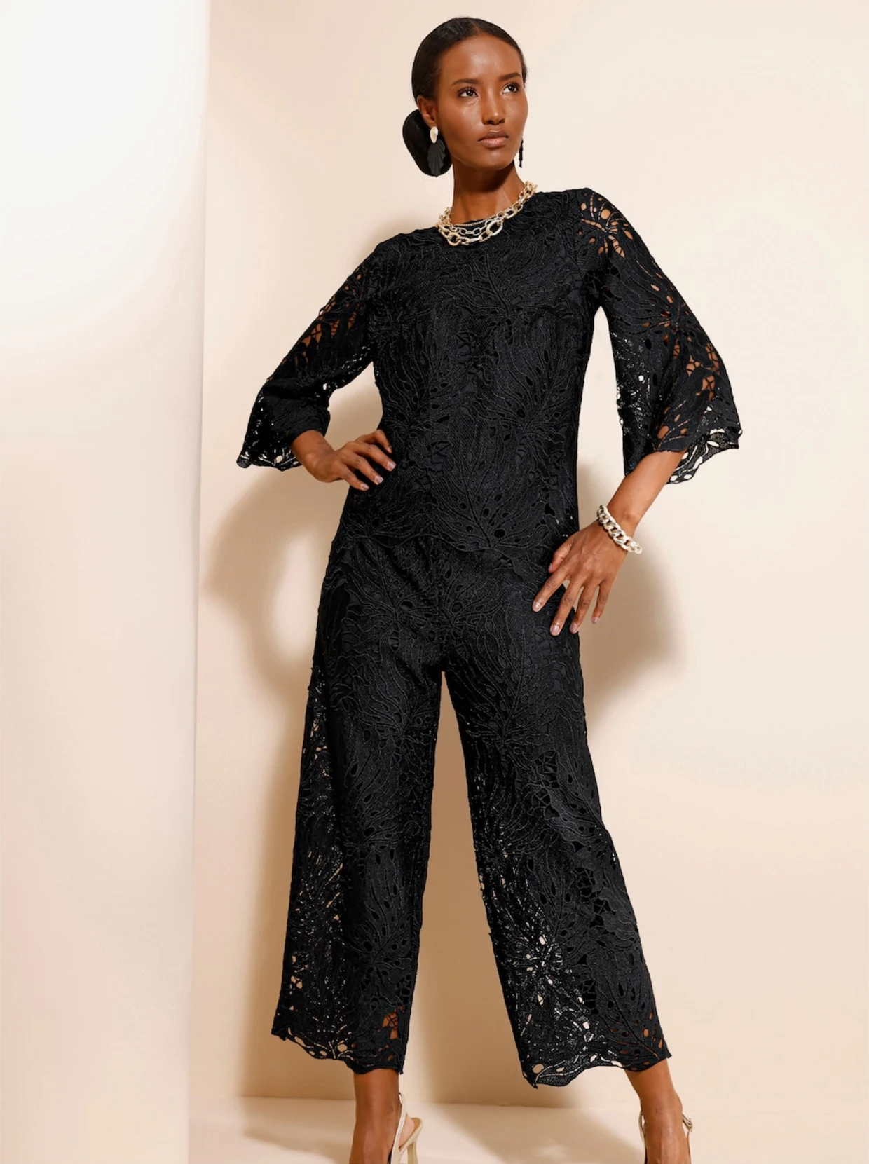 Weibliches Model stehend vor einer beigen Wand. Das Model trägt eine schwarze Spitzenbluse und dazu eine passende Hose. Weibliches Model stehend vor einer beigen Wand. Das Model trägt eine schwarze Spitzenbluse und dazu eine passende Hose.