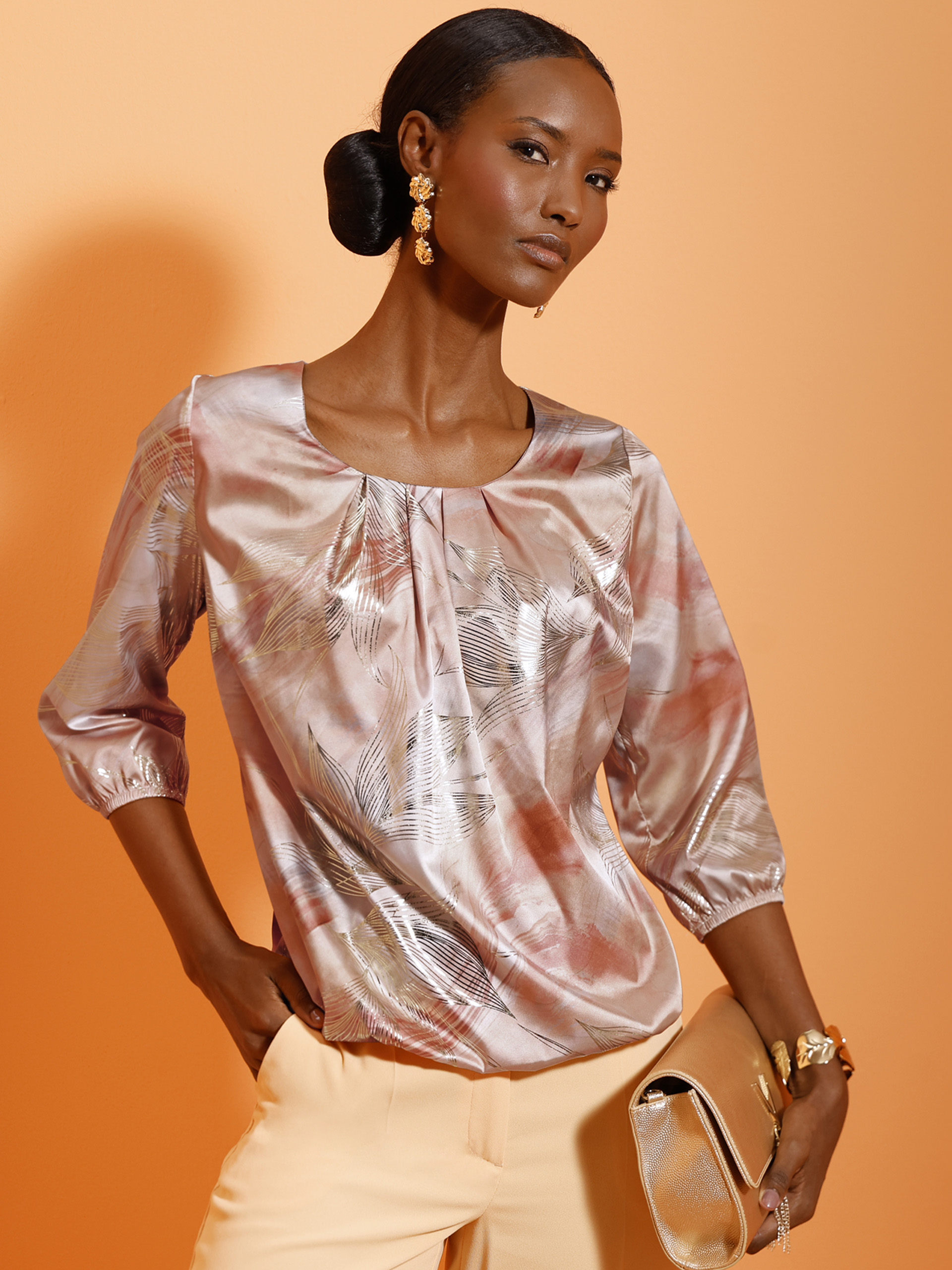 Ein Model mit hochgestecktem Haar trägt eine glänzende, gemusterte Bluse mit 3/4-Ärmeln und helle Hose, dazu goldene Accessoires.