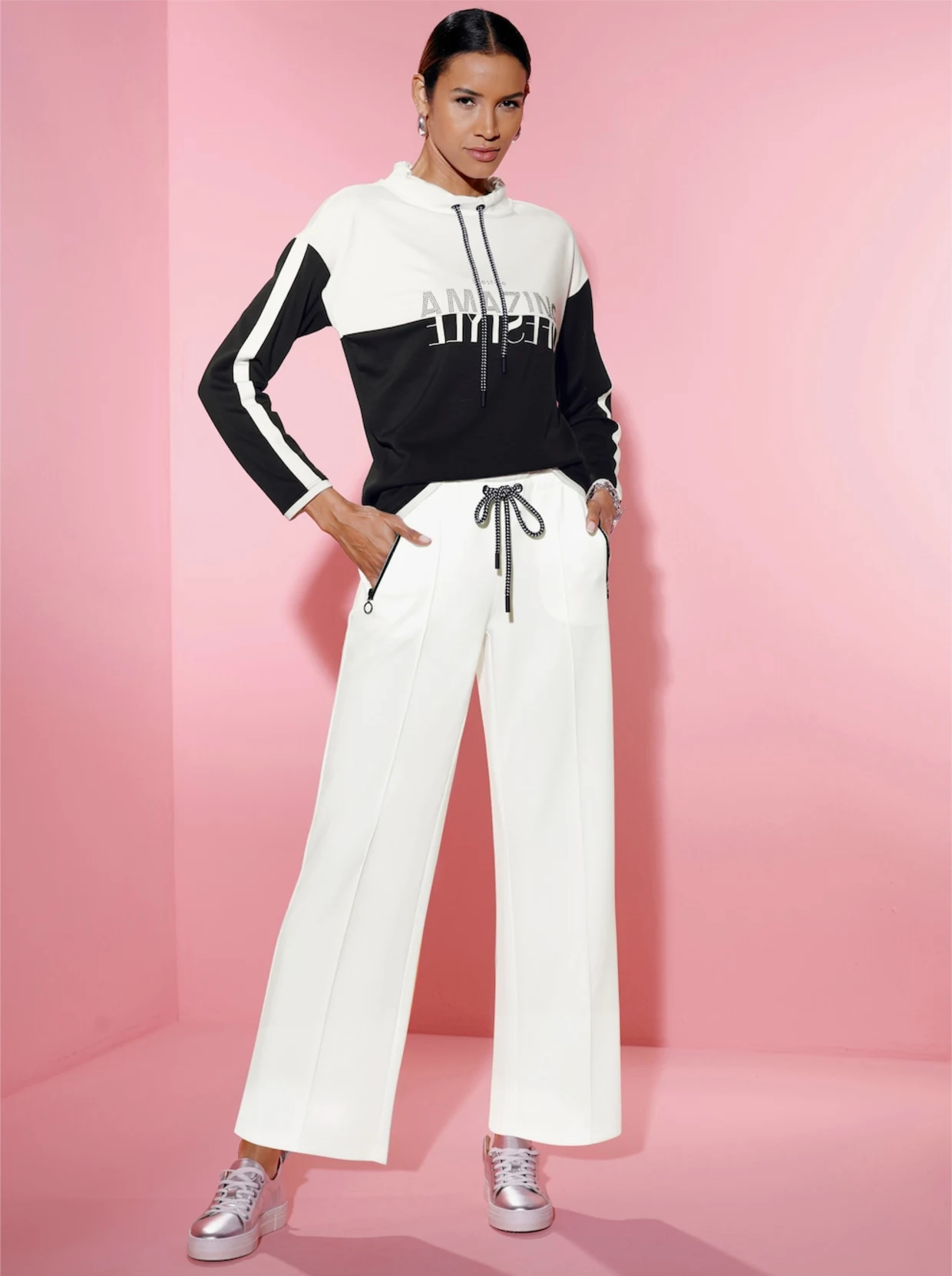 Weibliches Model stehend vor einem rosa Hintergrund. Das Model trägt eine weiße Hose und ein Sweatshirt mit schwarz weißen Blockstreifen. Weibliches Model stehend vor einem rosa Hintergrund. Das Model trägt eine weiße Hose und ein Sweatshirt mit schwarz weißen Blockstreifen.