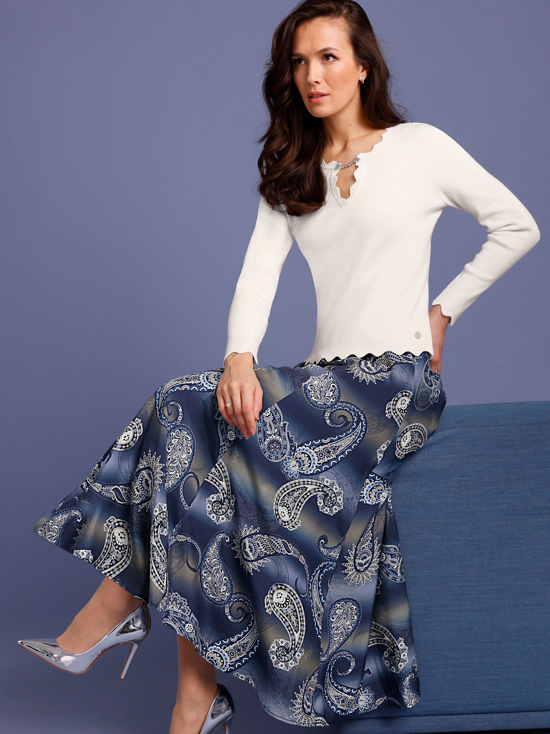 Ein Model sitzt auf einem blauen Möbelstück, trägt einen weißen Pullover und einen langen, blau gemusterten Rock mit Paisley-Design.