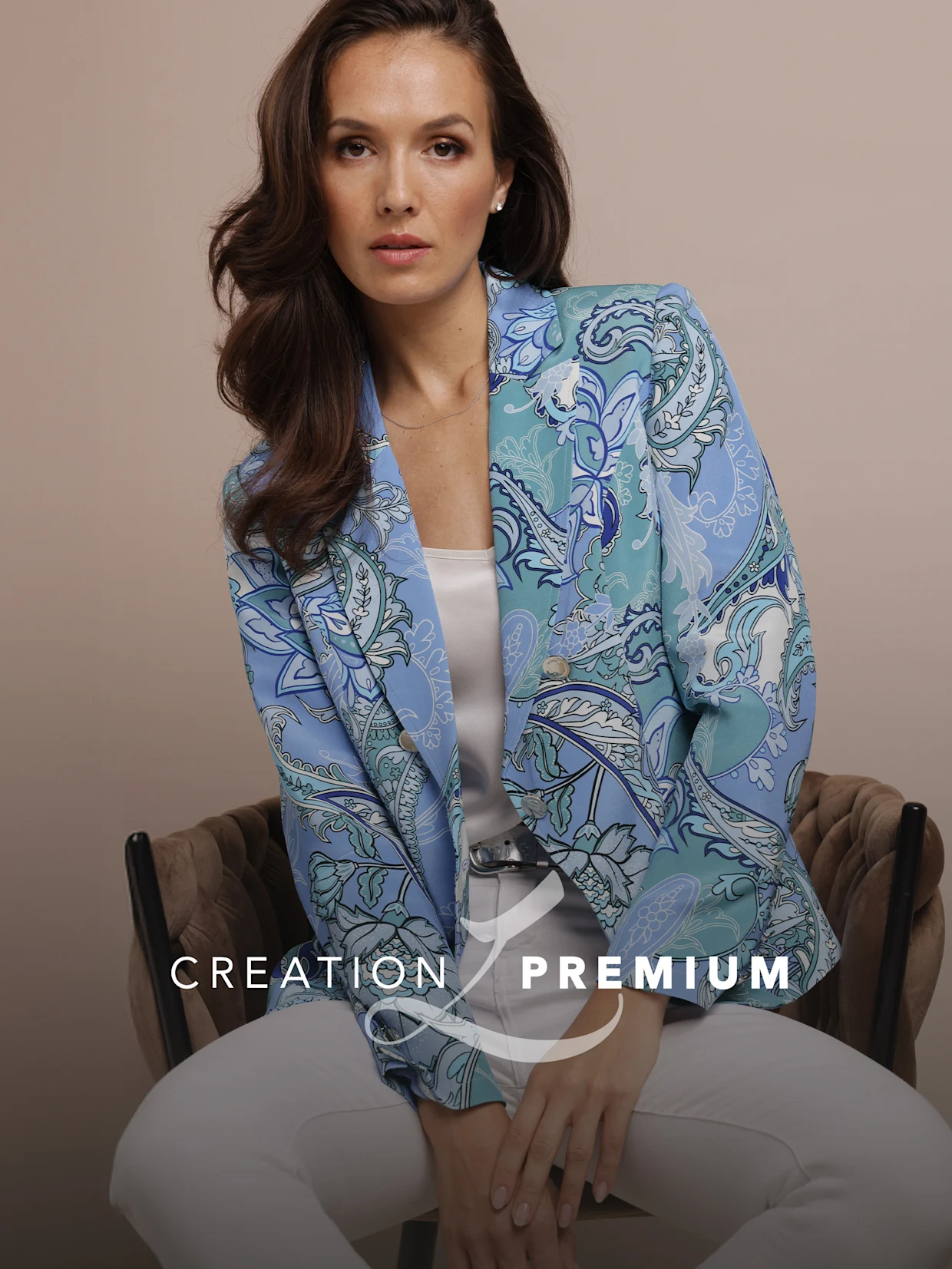 Ein Model sitzt auf einem Stuhl, trägt einen gemusterten Blazer in Blau- und Grüntönen zu weißer Hose. Text: CREATION PREMIUM.