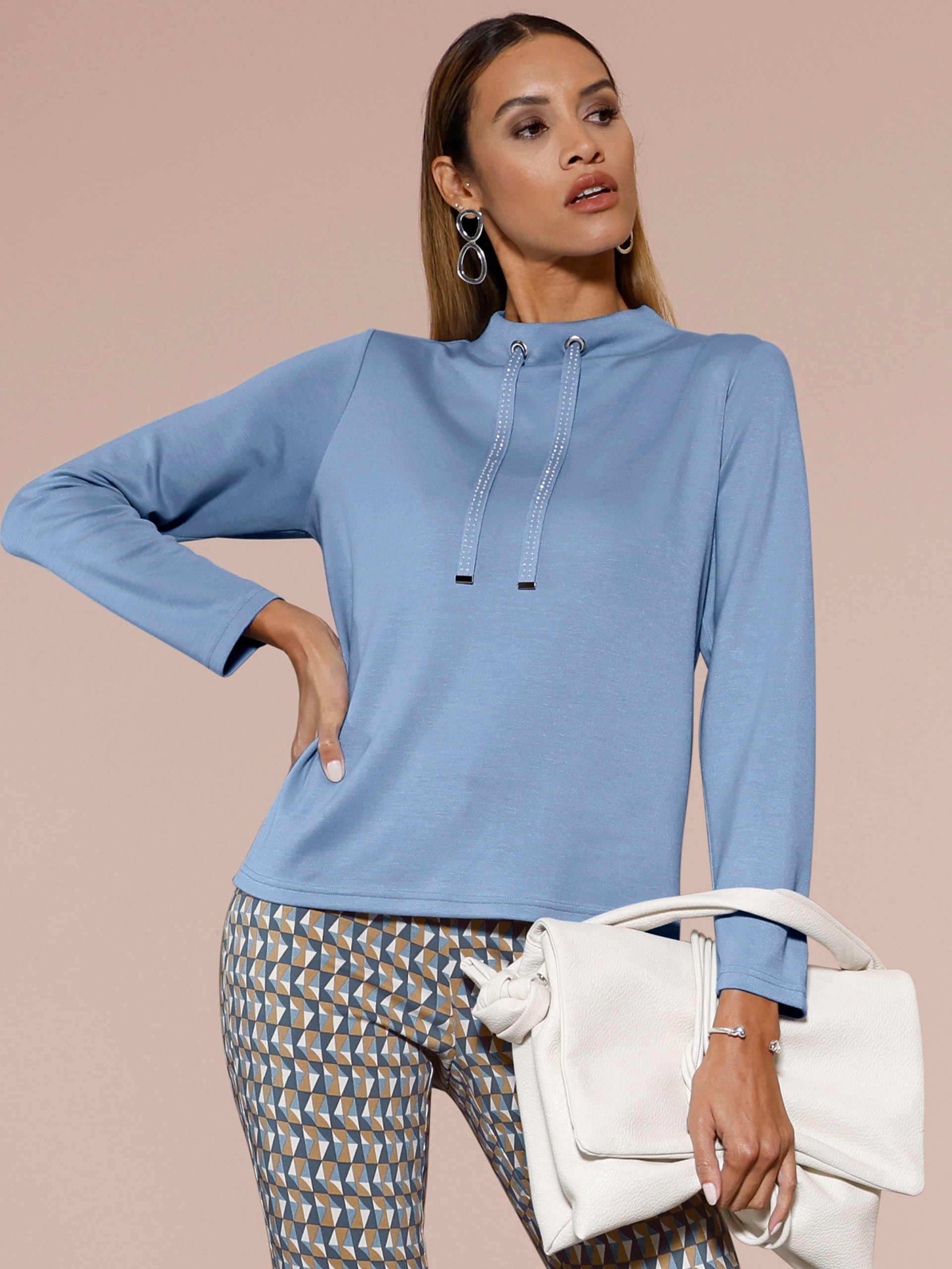 Ein Model trägt einen hellblauen Pullover mit Tunnelzug am Kragen, dazu gemusterte Hose und auffällige Ohrringe.