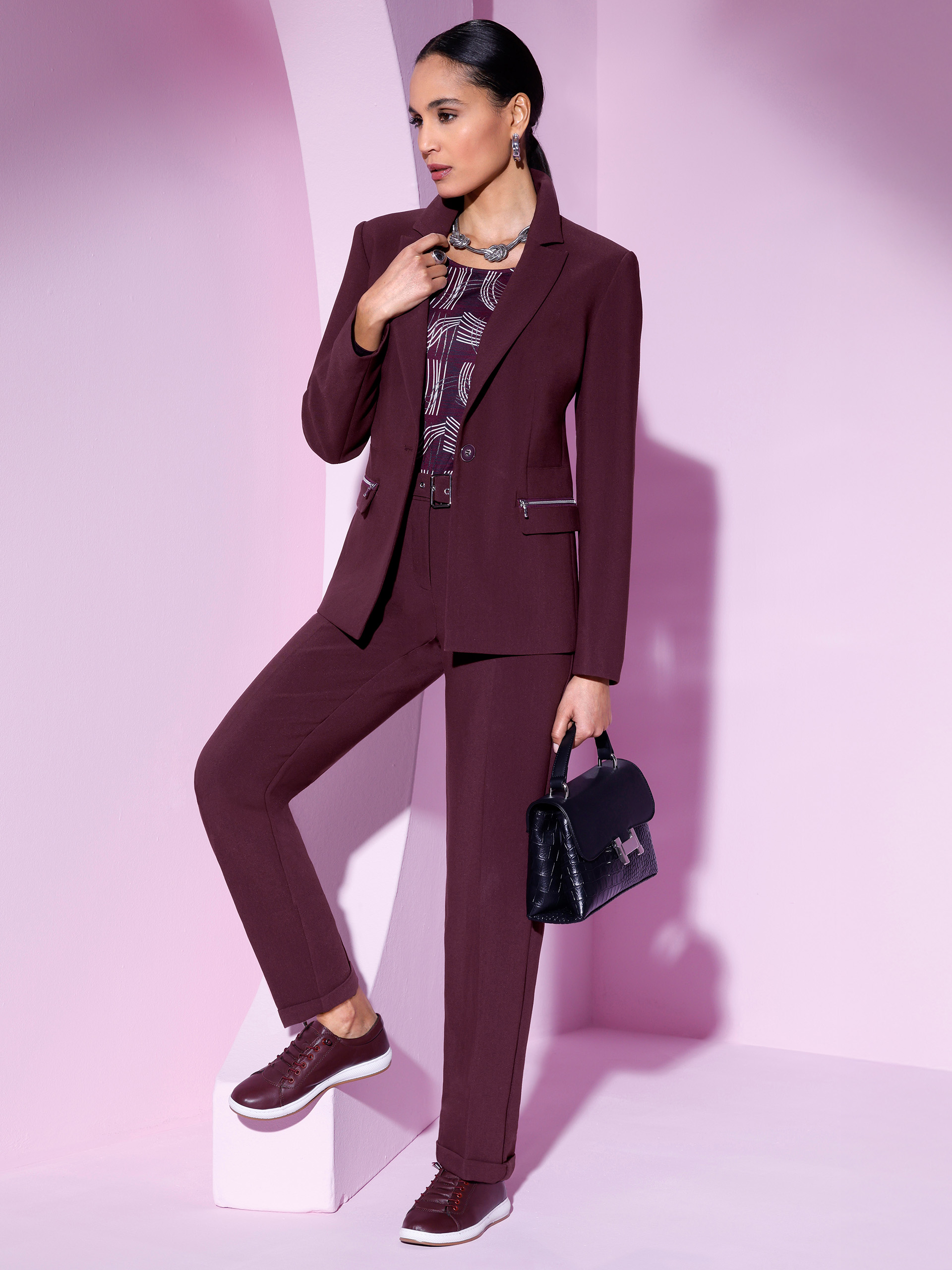 Ein Model trägt einen burgunderfarbenen Anzug mit einer gemusterten Bluse und sportlichen Schuhen. Sie hält eine schwarze Handtasche. Der Hintergrund ist rosa.