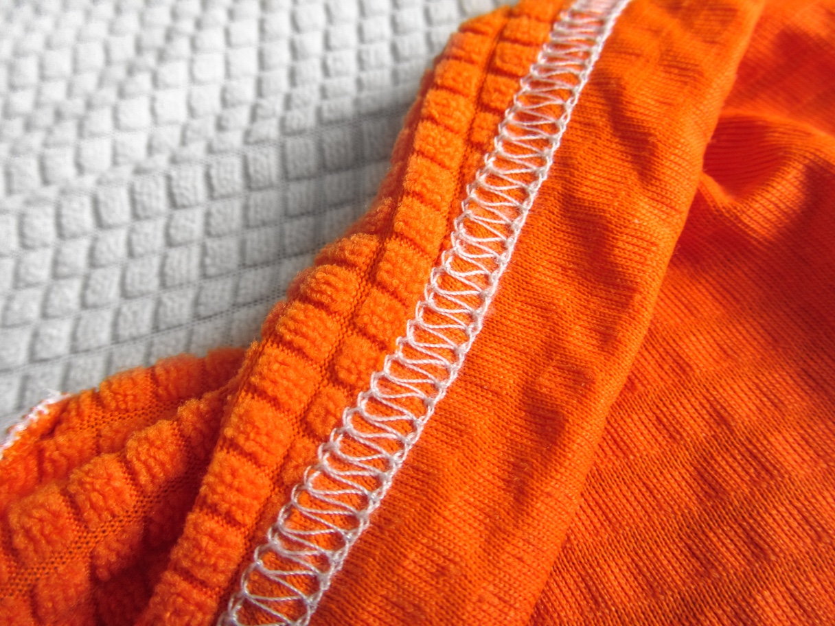 Nahaufnahme eines orangefarbenen Stoffes mit weißer Naht und strukturierter Oberfläche, liegend auf einem weißen, texturierten Hintergrund.