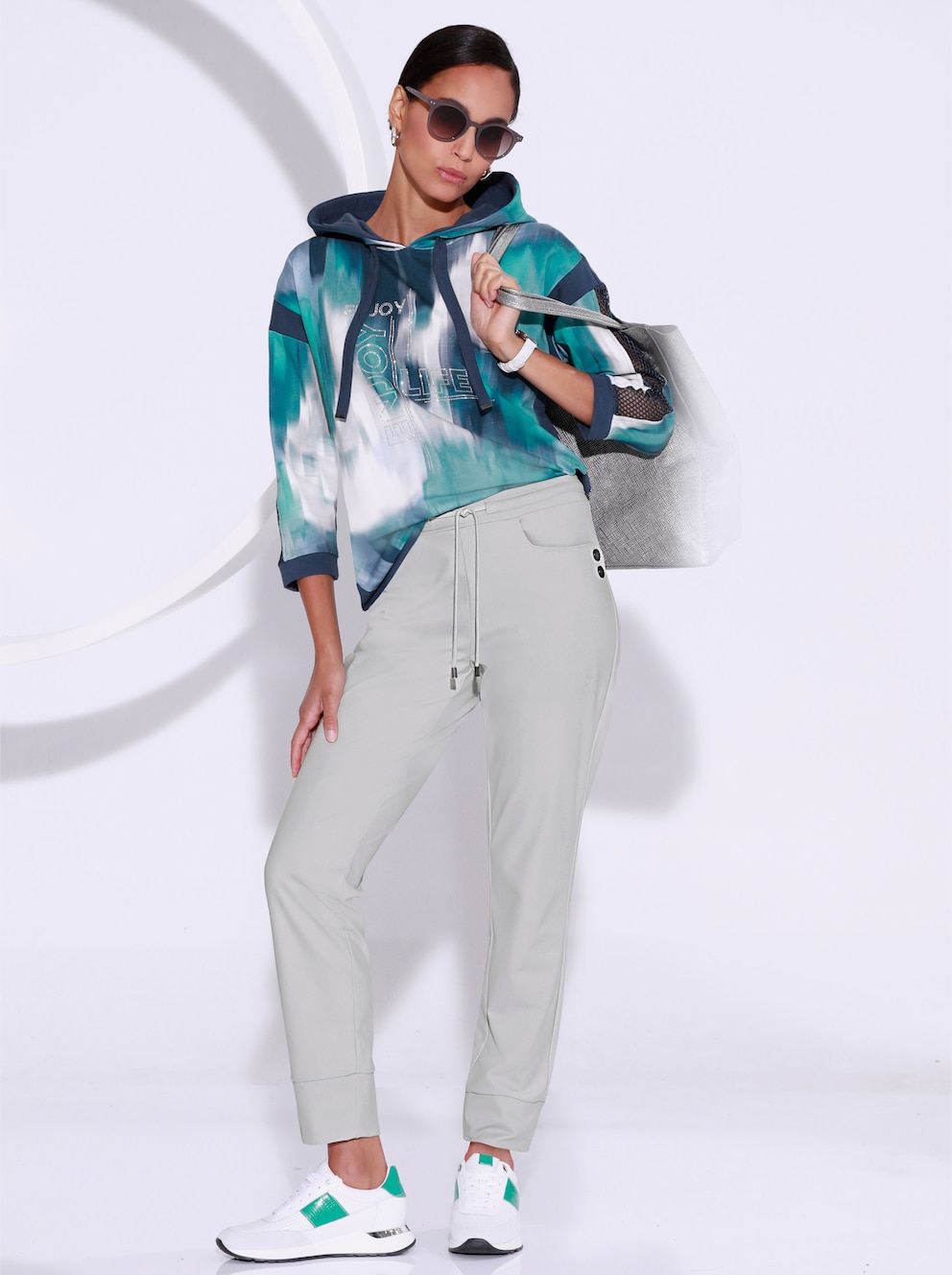 Weibliches Model stehende vor einer weißen Fotoleinwand. Das Model trägt eine graue Hose, ein Sweatshirt in den Farben salbei-dunkelblau und weiße Sneaker.