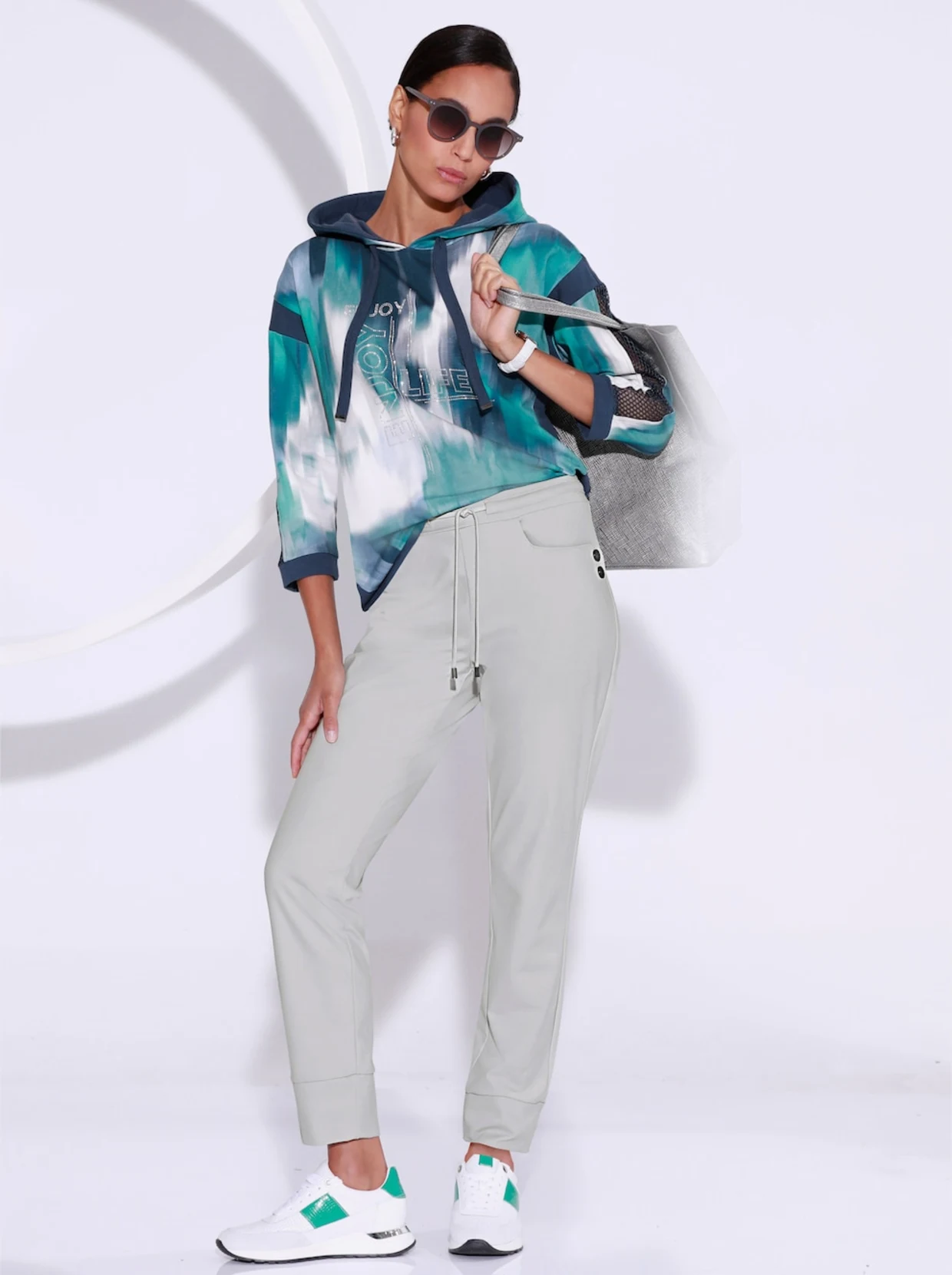 Weibliches Model stehende vor einer weißen Fotoleinwand. Das Model trägt eine graue Hose, ein Sweatshirt in den Farben salbei-dunkelblau und weiße Sneaker. Weibliches Model stehende vor einer weißen Fotoleinwand. Das Model trägt eine graue Hose, ein Sweatshirt in den Farben salbei-dunkelblau und weiße Sneaker.