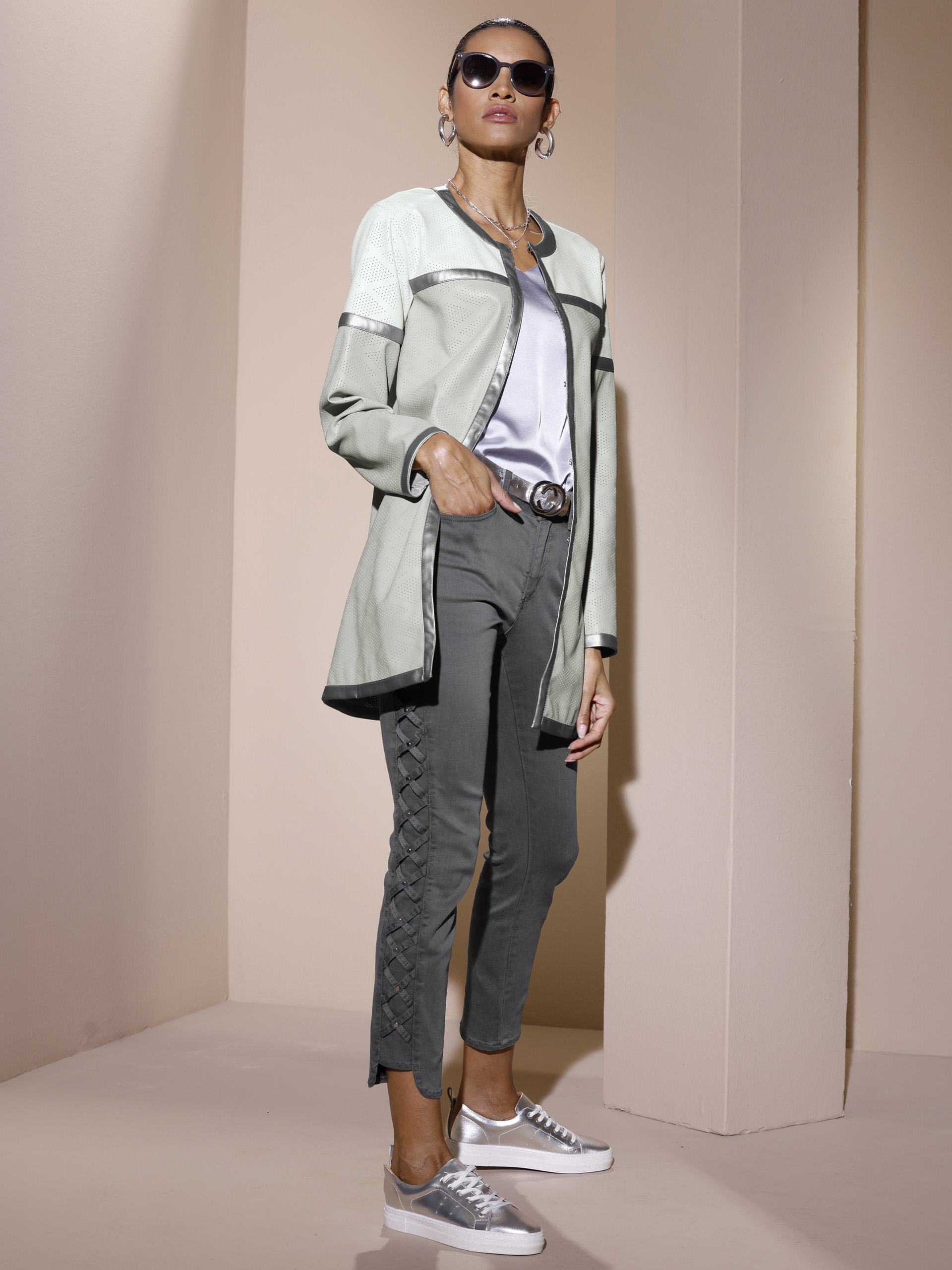 Ein Model steht vor beigefarbenem Hintergrund, trägt eine lange hellgraue Jacke, ein weißes Shirt, graue Hose mit Schnürdetails und silberne Sneaker.