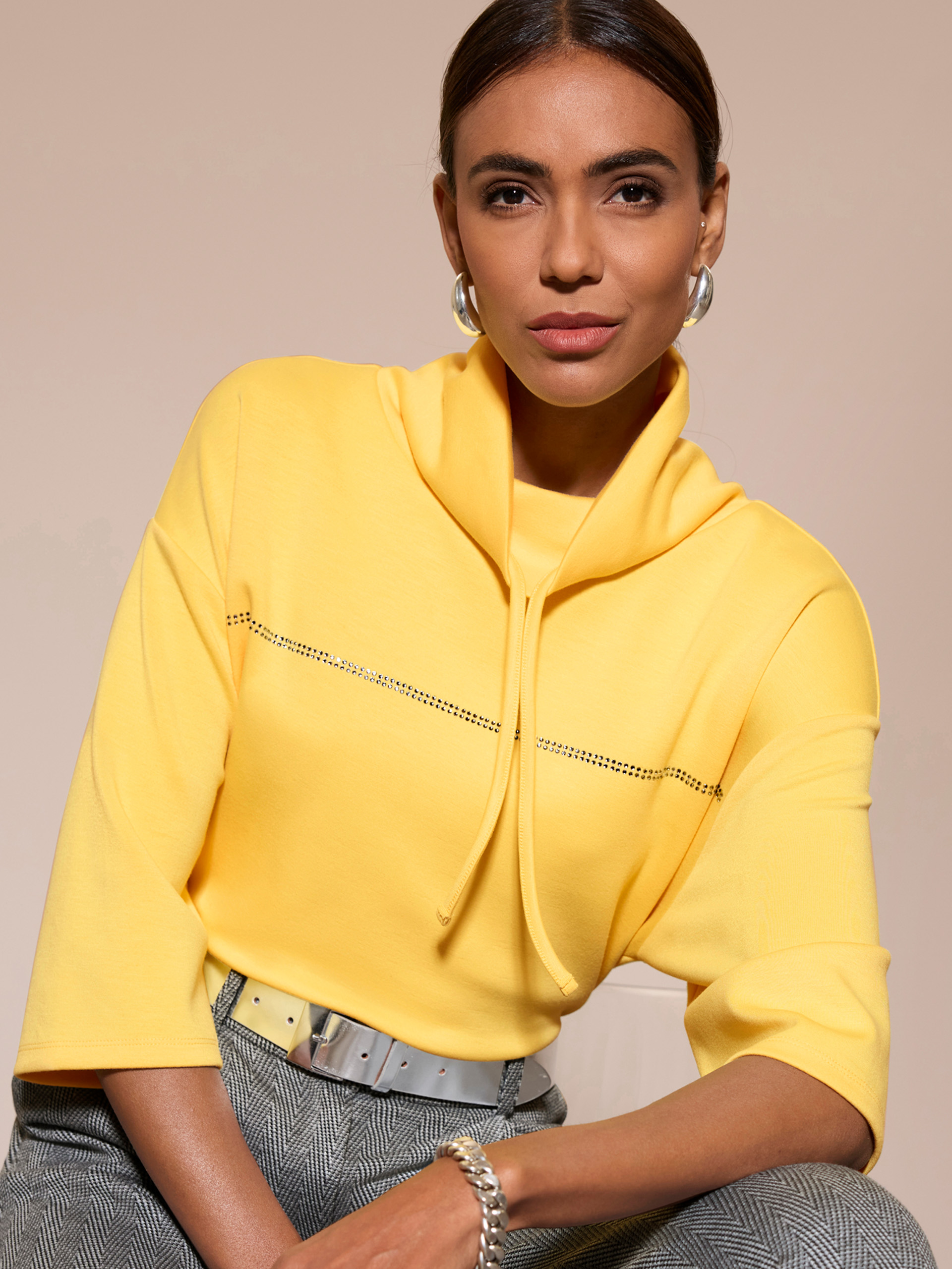 Ein Model trägt einen gelben Pullover mit Stehkragen und kurzen Ärmeln, dazu eine graue Hose und silbernen Gürtel.