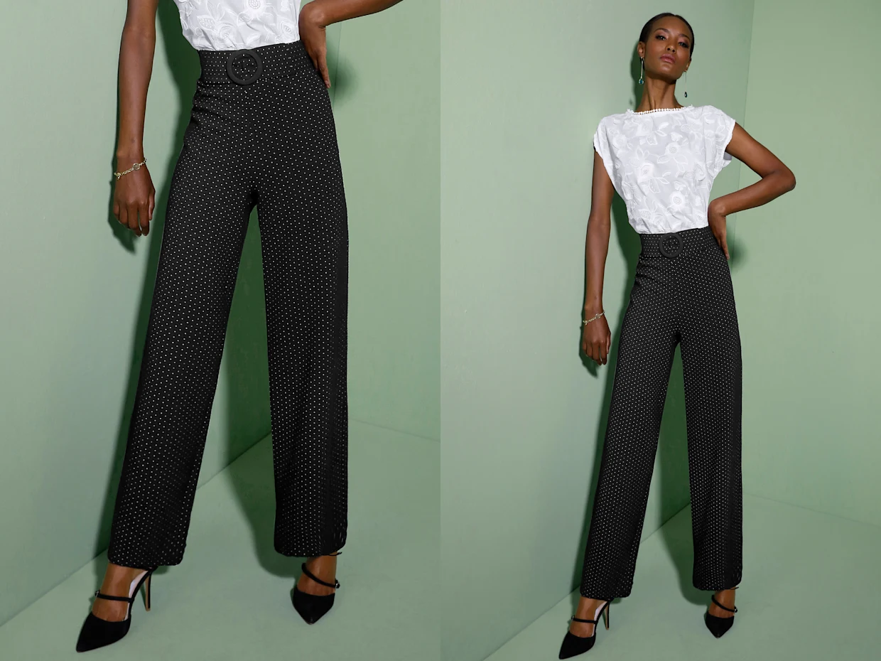 Schwarze, weit geschnittene Hose mit weißen Punkten und hoher Taille, kombiniert mit weißem Oberteil mit floralen Stickereien, getragen mit schwarzen Absatzschuhen vor grünem Hintergrund.