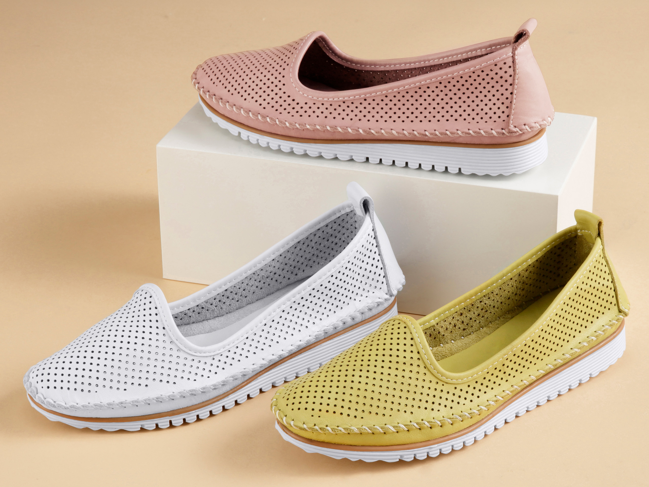 Drei Paar Slipper-Schuhe in Rosa, Weiß und Gelb mit Lochmuster und weißer Sohle auf hellem Untergrund arrangiert.