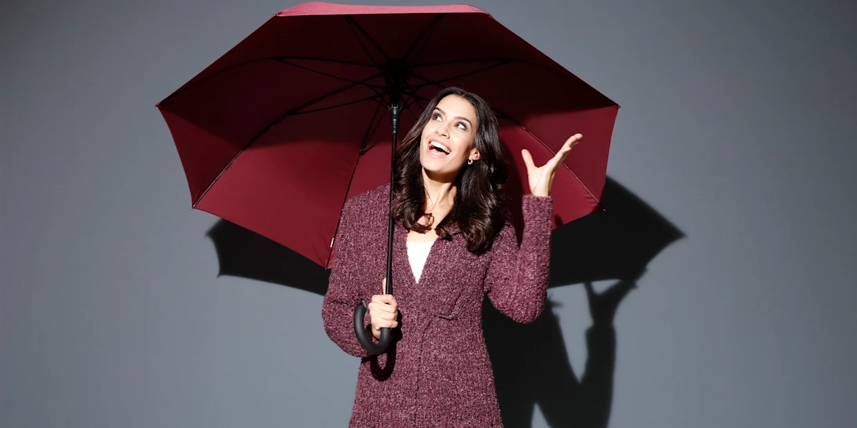 Ein Model hält einen roten Regenschirm und gestikuliert mit der anderen Hand vor einem grauen Hintergrund. Ein Model hält einen roten Regenschirm und gestikuliert mit der anderen Hand vor einem grauen Hintergrund.