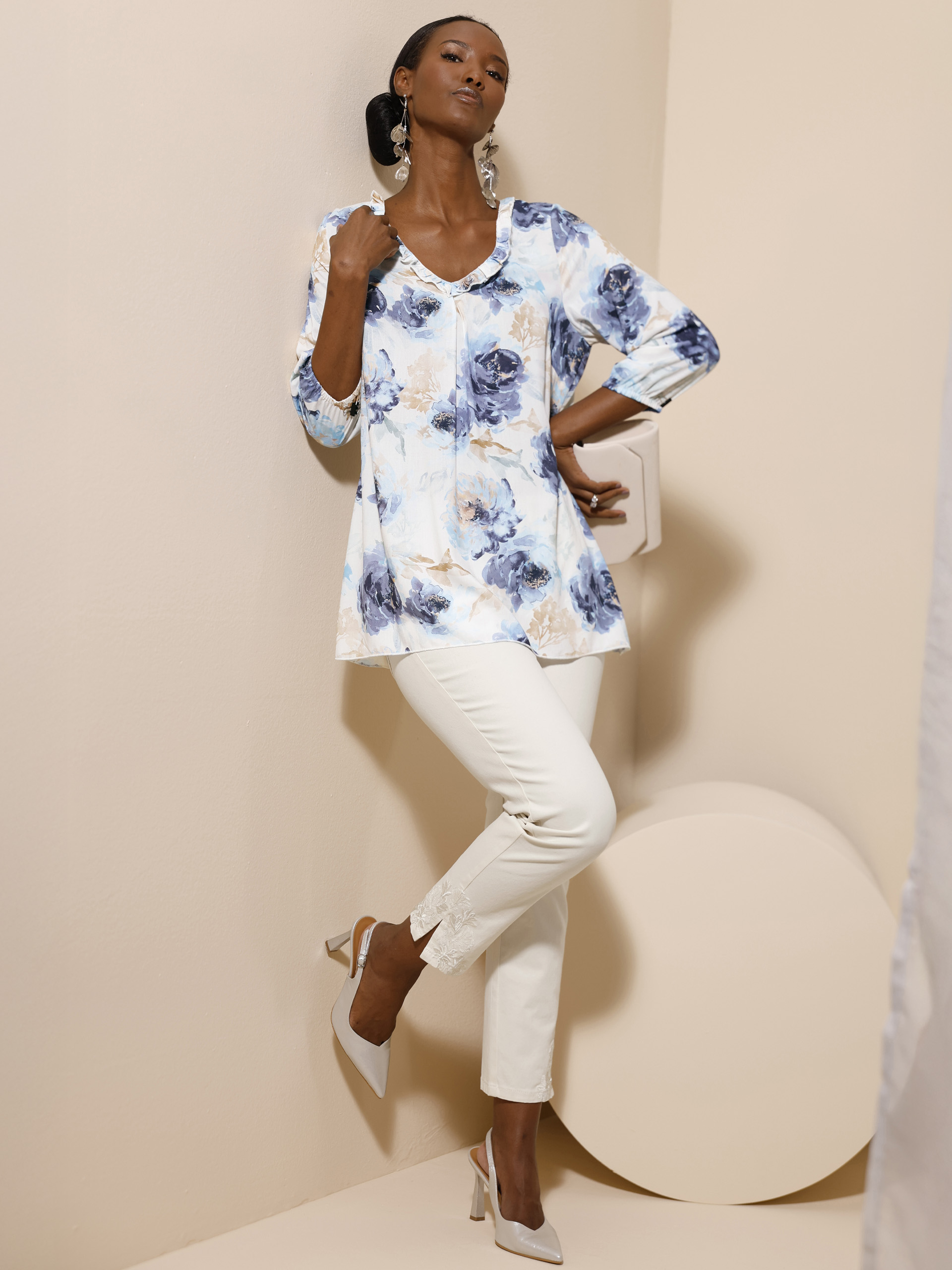 Ein Model steht vor beigem Hintergrund, trägt eine weiße Hose mit Spitzenbesatz und eine geblümte Tunika in Blau- und Beigetönen sowie helle Pumps.