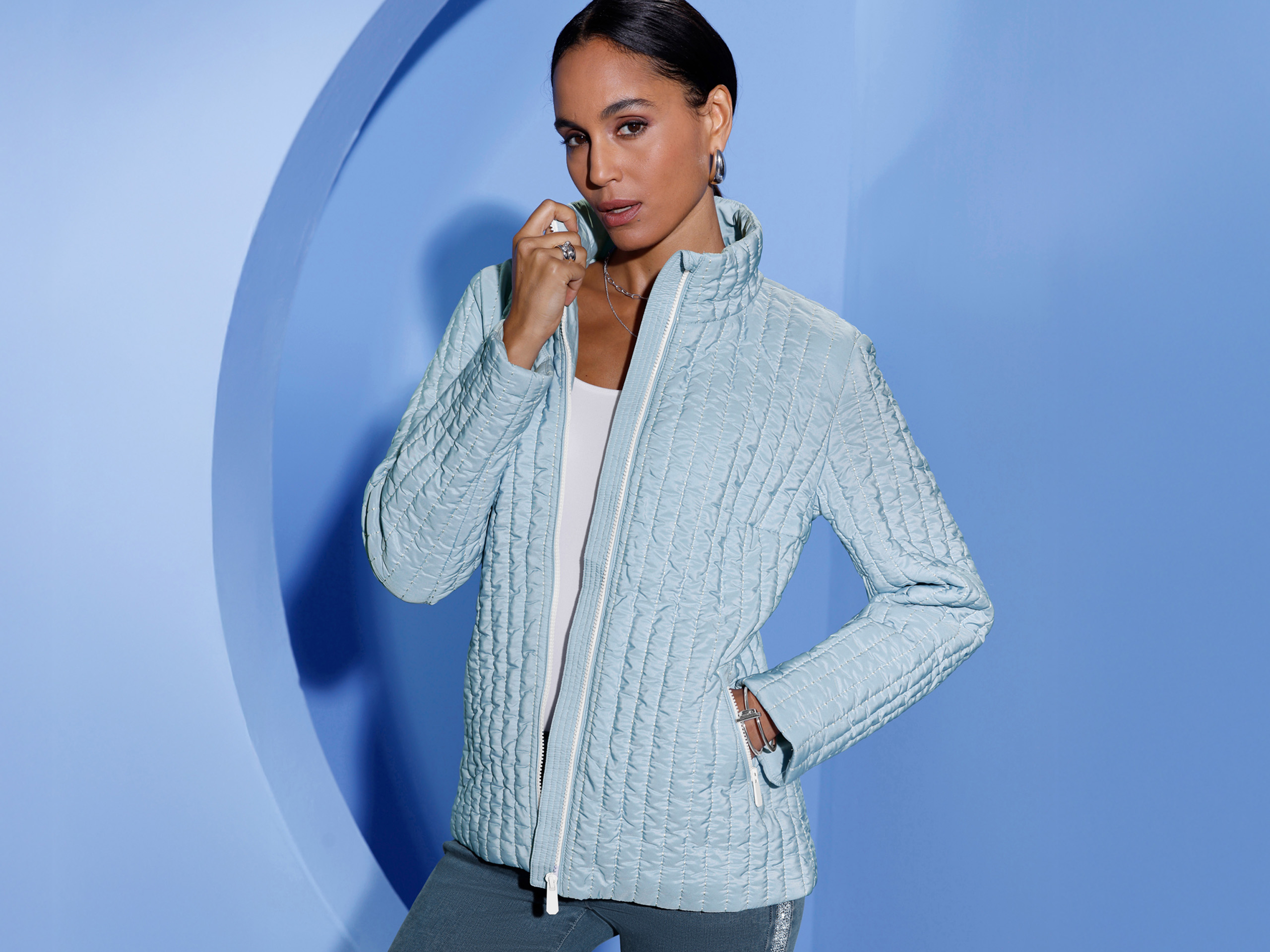 Ein Model trägt eine hellblaue, gesteppte Jacke mit Reißverschluss vor einem hellblauen Hintergrund.