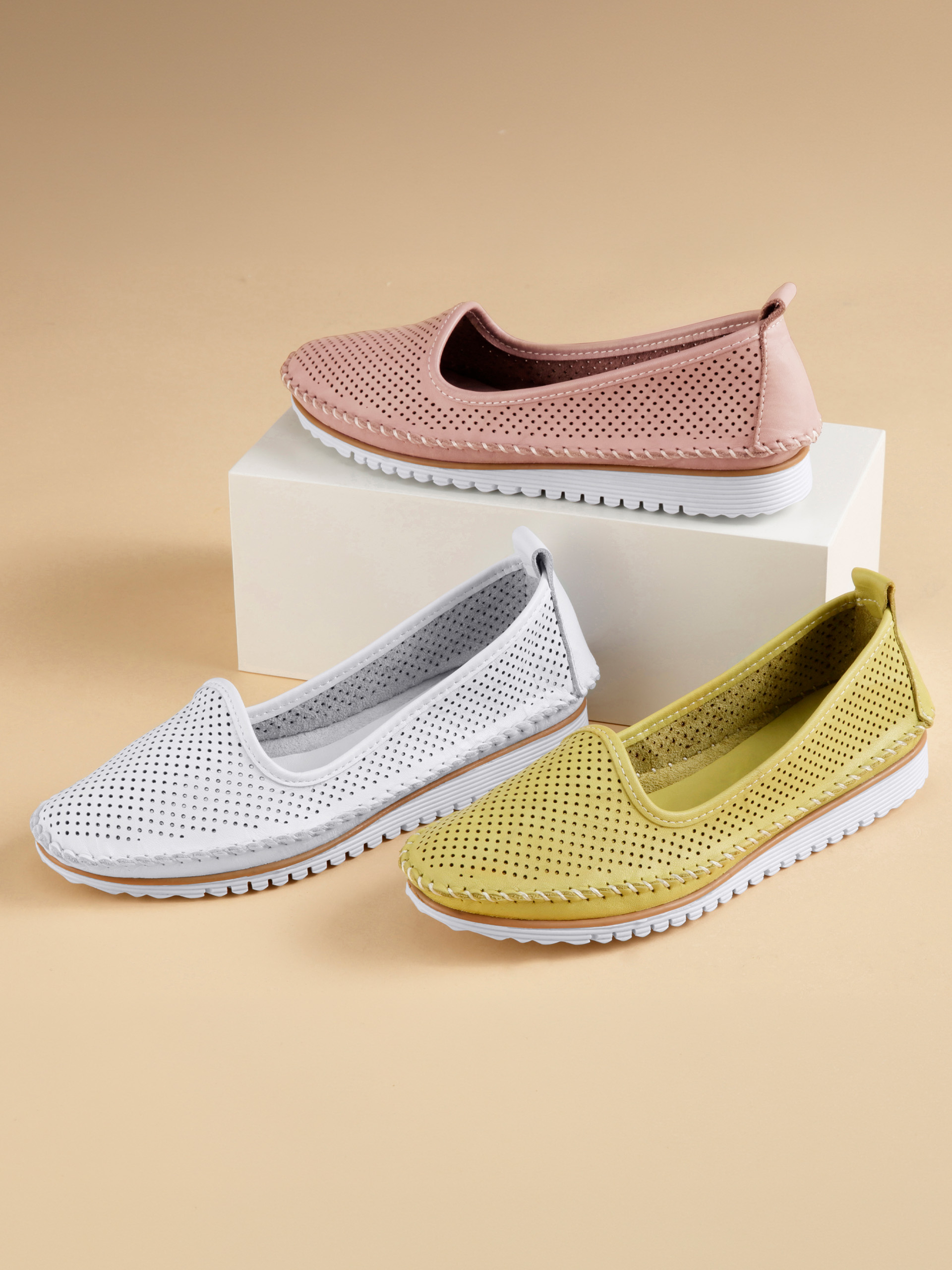 Drei Paar Slipper-Schuhe in Rosa, Weiß und Gelb mit Lochmuster und weißer Sohle auf hellem Untergrund arrangiert.