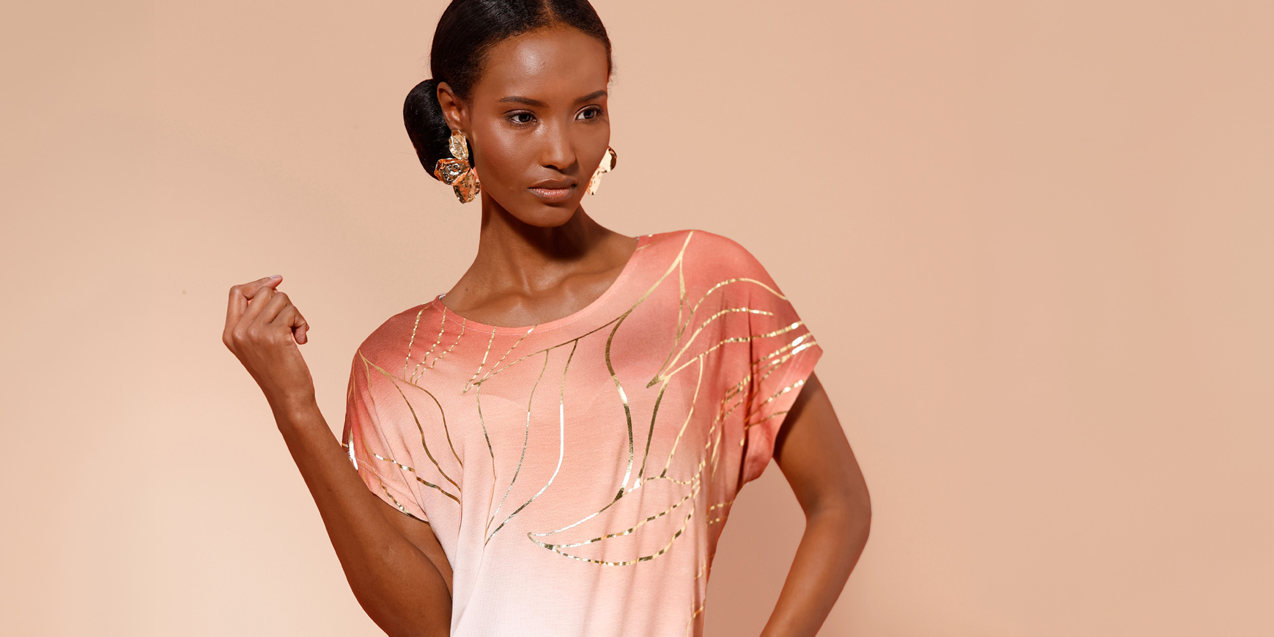 Model in apricotfarbenem Shirt mit goldenen Akzenten vor beigem Hintergrund.