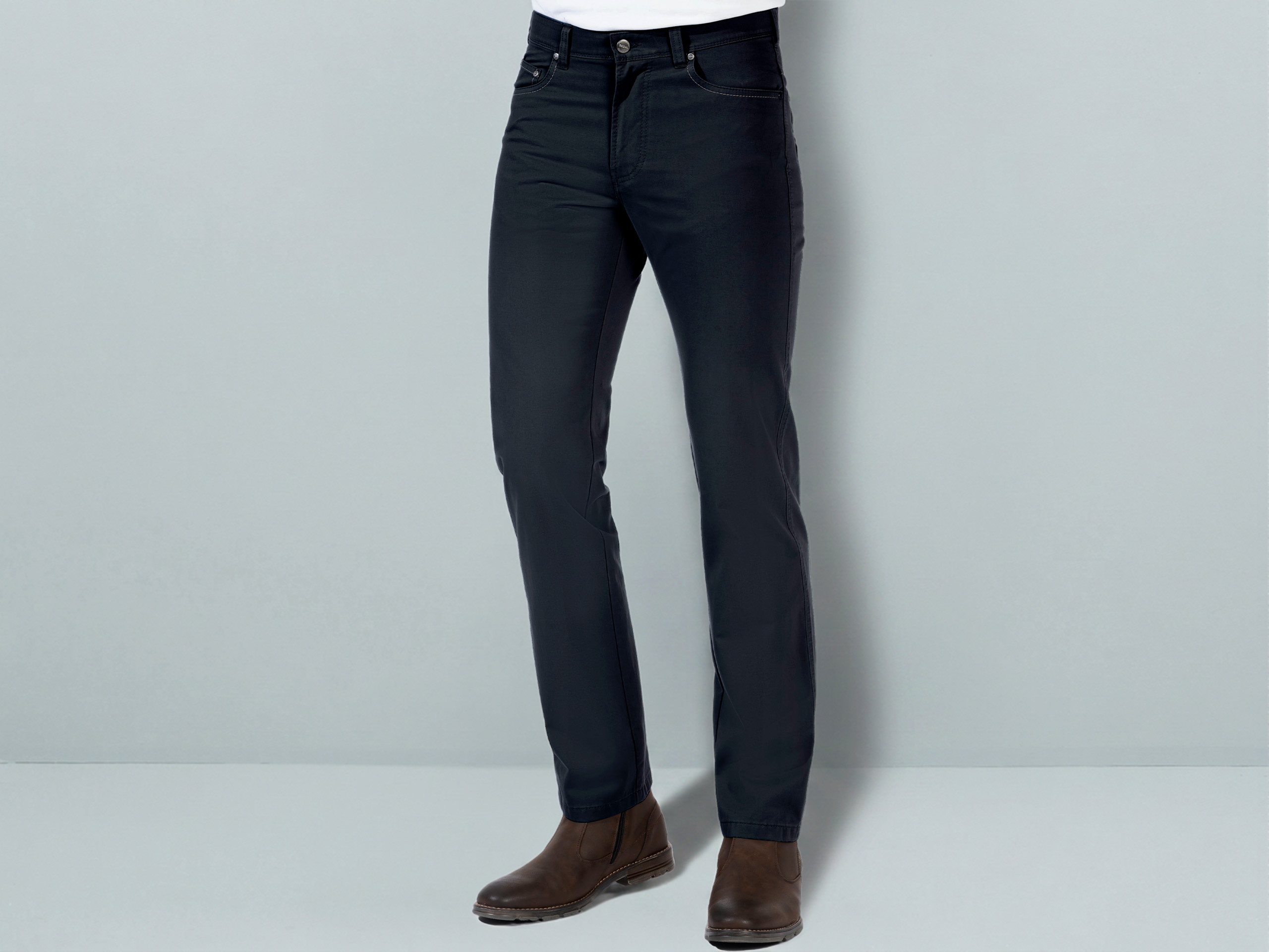 Mann mit dunkler Jeans und braunen Schuhen vor grauem Hintergrund.