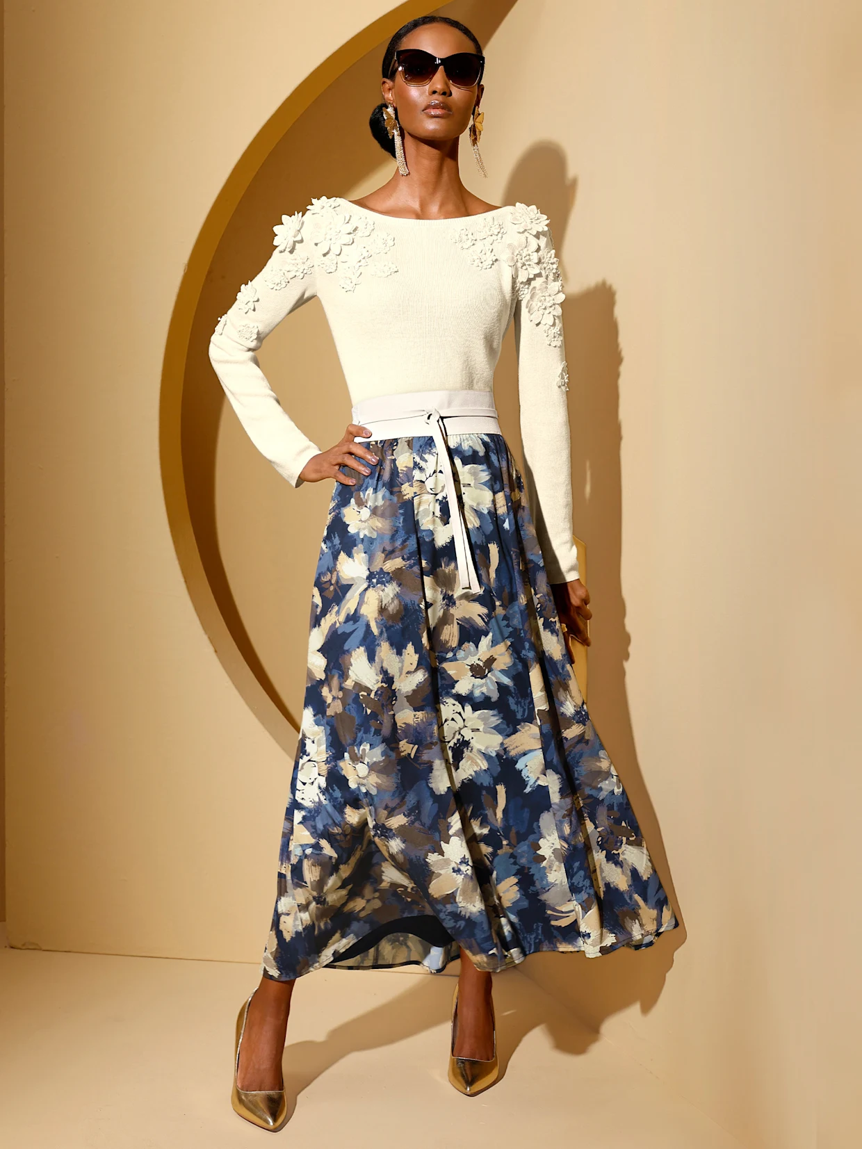 Ein Model trägt ein weißes Oberteil mit floralen Details an den Schultern und langen Ärmeln, kombiniert mit einem langen, blauen Rock mit floralen Mustern. Die Schuhe sind hoch und elegant. Der Hintergrund ist in warmen Tönen gehalten. Ein Model trägt ein weißes Oberteil mit floralen Details an den Schultern und langen Ärmeln, kombiniert mit einem langen, blauen Rock mit floralen Mustern. Die Schuhe sind hoch und elegant. Der Hintergrund ist in warmen Tönen gehalten.