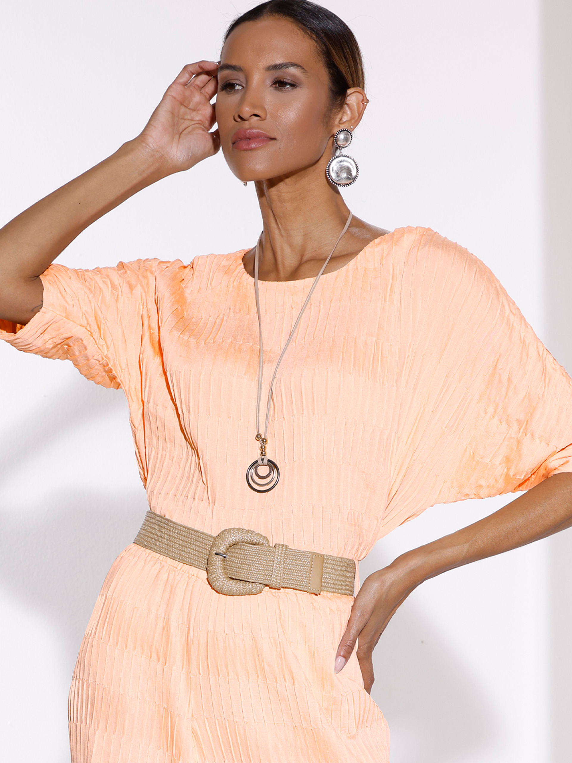 Ein Model trägt ein apricotfarbenes, strukturiertes Kleid mit breitem Gürtel, auffälligen Ohrringen und langer Kette vor weißem Hintergrund.