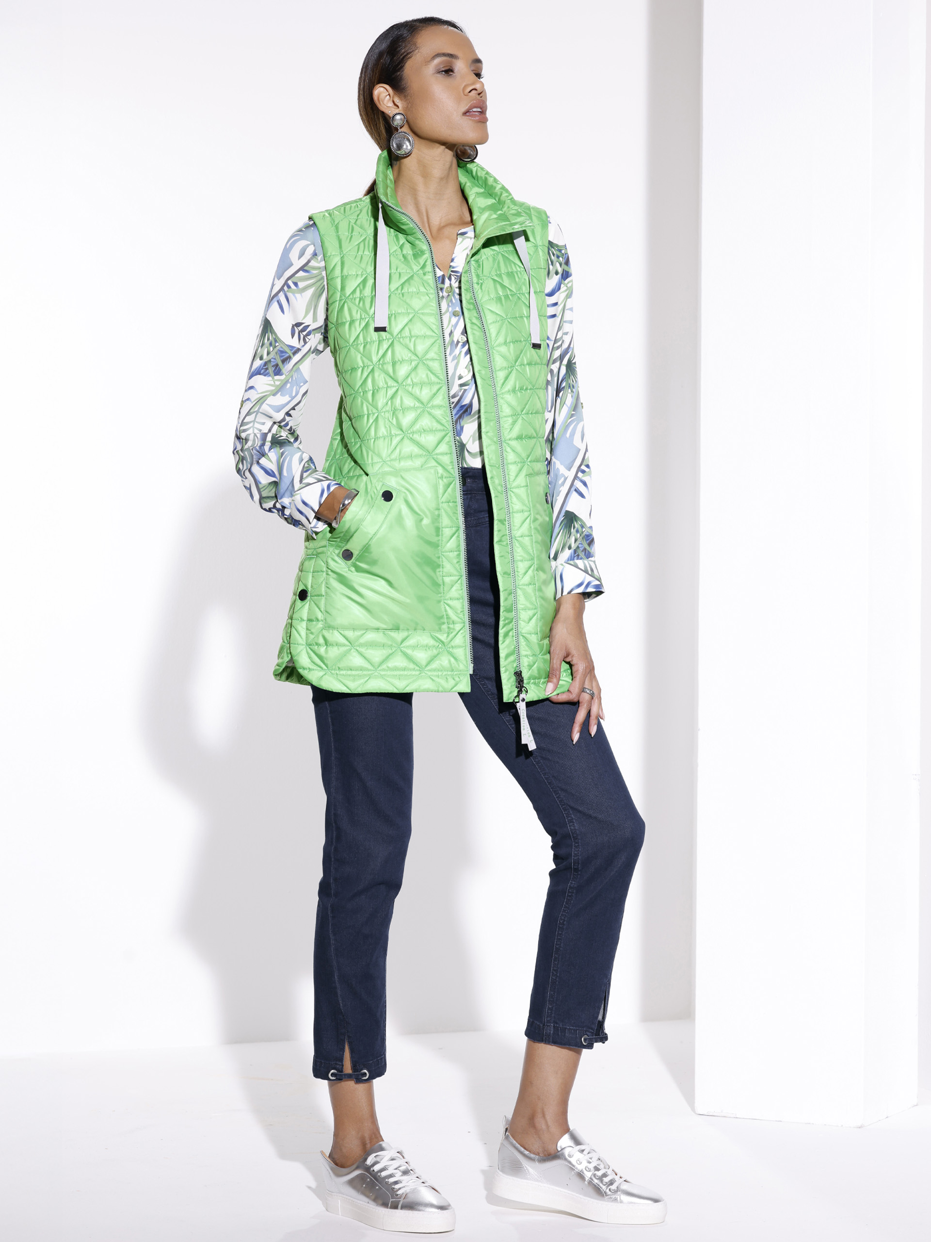 Ein Model steht vor weißem Hintergrund, trägt eine grüne Steppweste, gemusterte Bluse, dunkelblaue Hose und helle Sneaker.