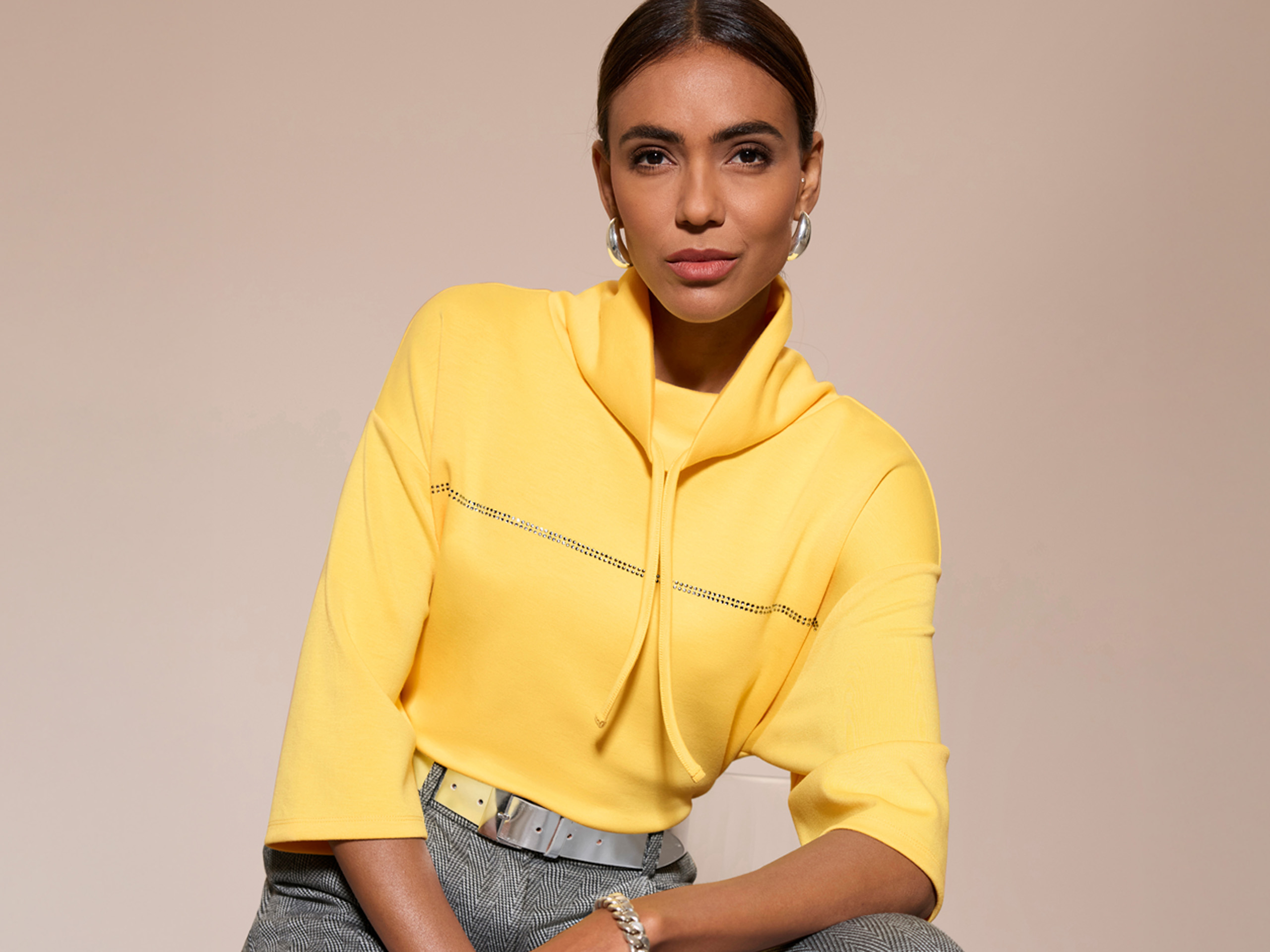Ein Model trägt einen gelben Pullover mit Stehkragen und kurzen Ärmeln, dazu eine graue Hose und silbernen Gürtel.