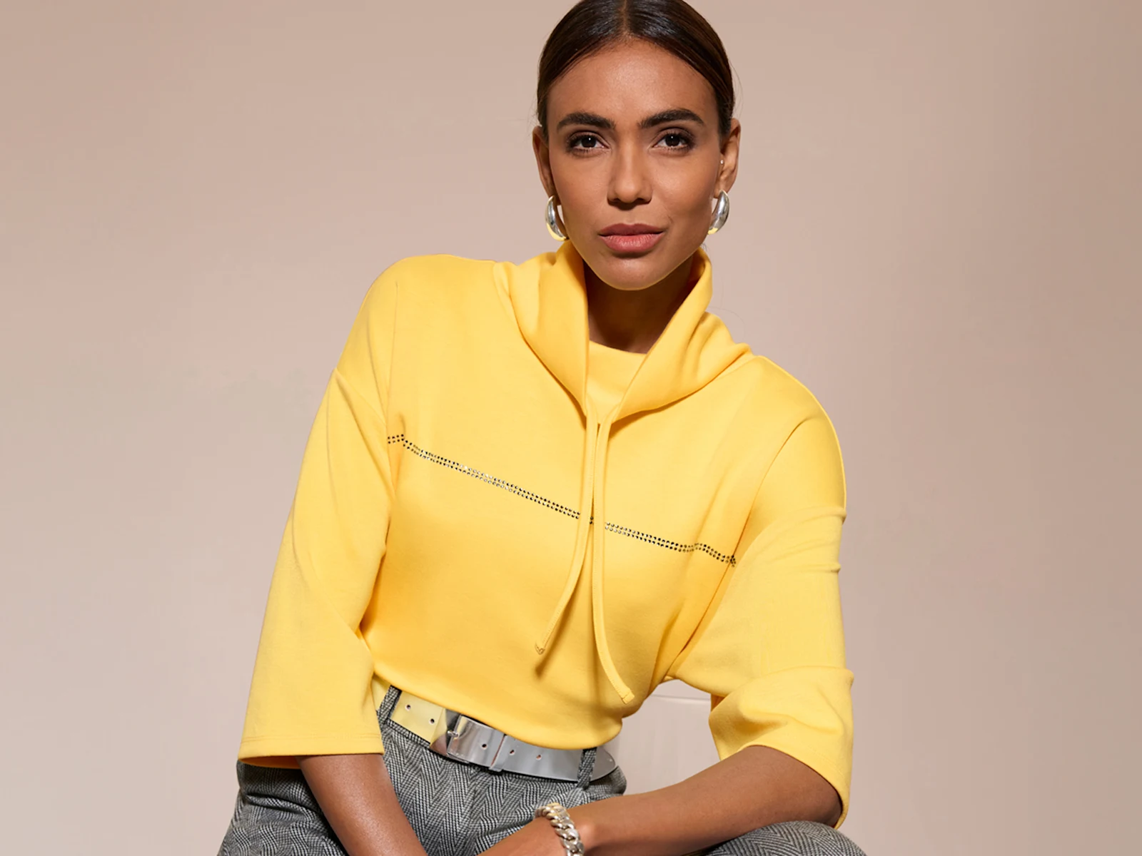 Ein Model trägt einen gelben Pullover mit Stehkragen und kurzen Ärmeln, dazu eine graue Hose und silbernen Gürtel.