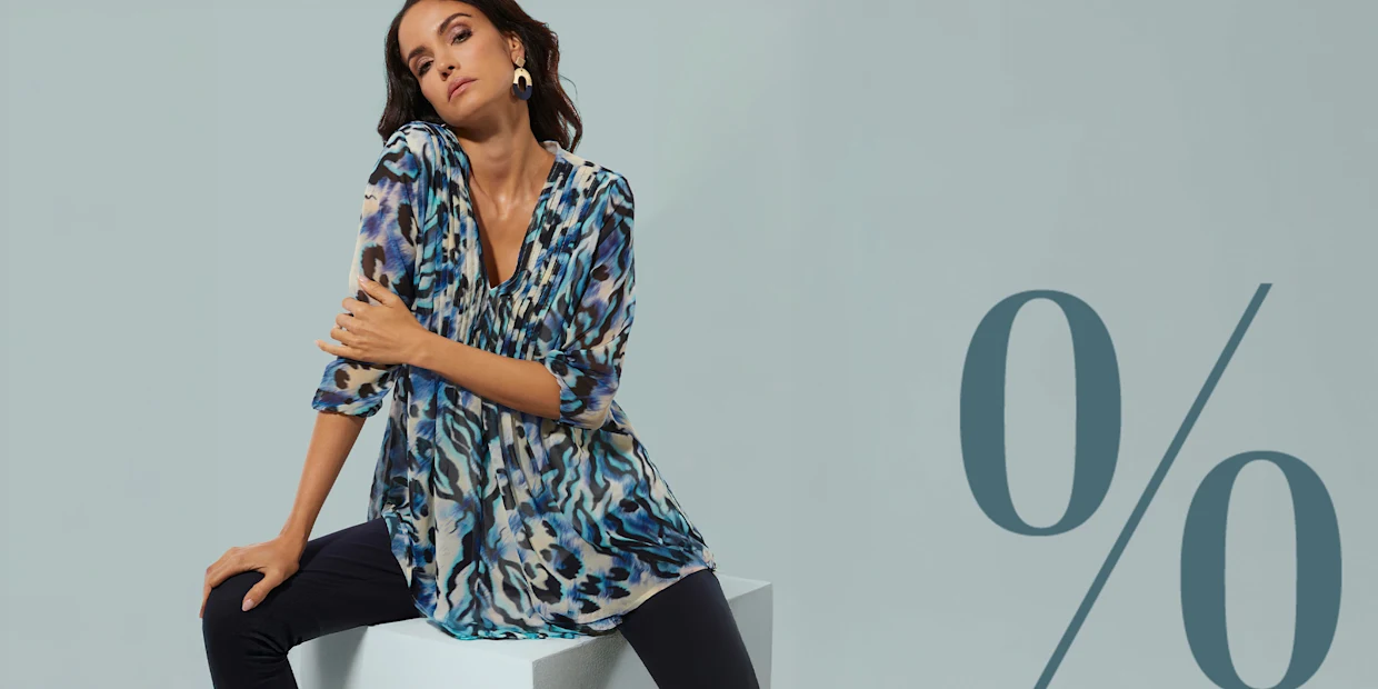 Ein Model sitzt auf einem Würfel, trägt eine gemusterte Tunika in Blau- und Beigetönen sowie dunkle Hose. Großes Prozentzeichen im Hintergrund.