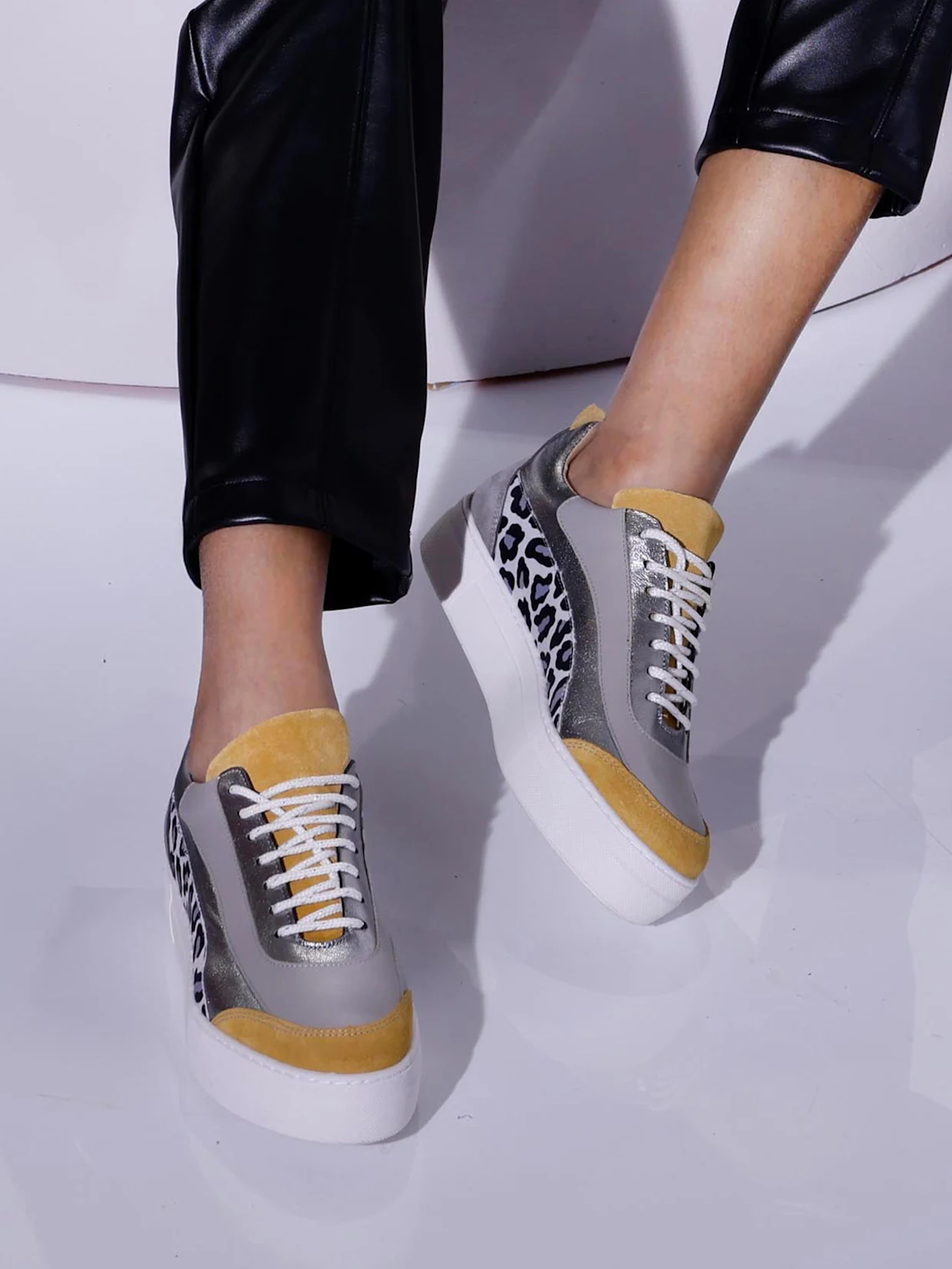 Ein Model trägt graue, gelbe und silberne Sneaker mit Leopardenmuster an den Seiten, dazu schwarze Lederhosen.