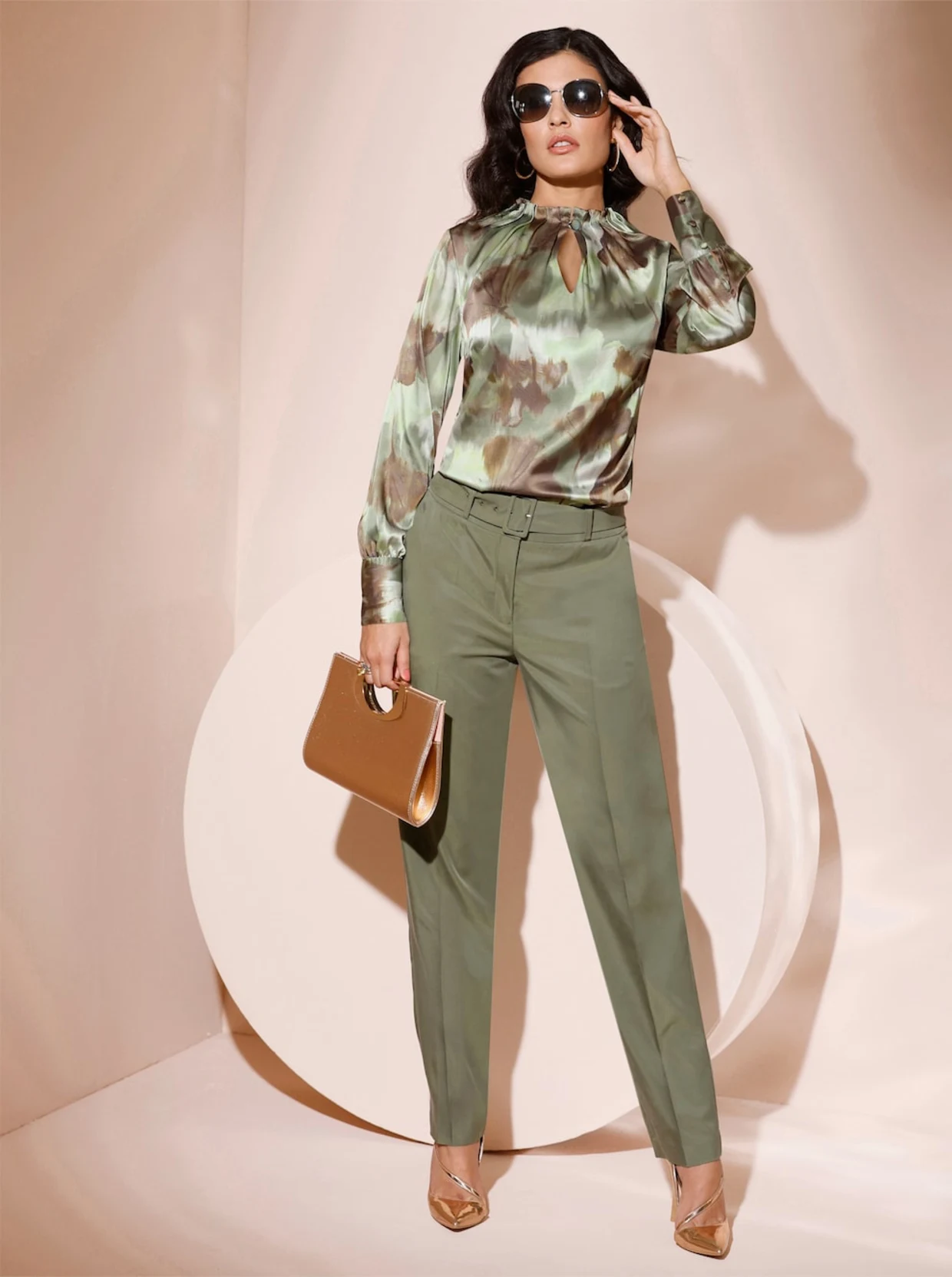 Weibliches Model stehen vor einer beigen Wand. Sie trägt eine Olive Farben Hose, dazu eine Satinbluse mit breiten Knopfmanschetten. In der Hand hält das Model eine braune Tasche. Weibliches Model stehen vor einer beigen Wand. Sie trägt eine Olive Farben Hose, dazu eine Satinbluse mit breiten Knopfmanschetten. In der Hand hält das Model eine braune Tasche.