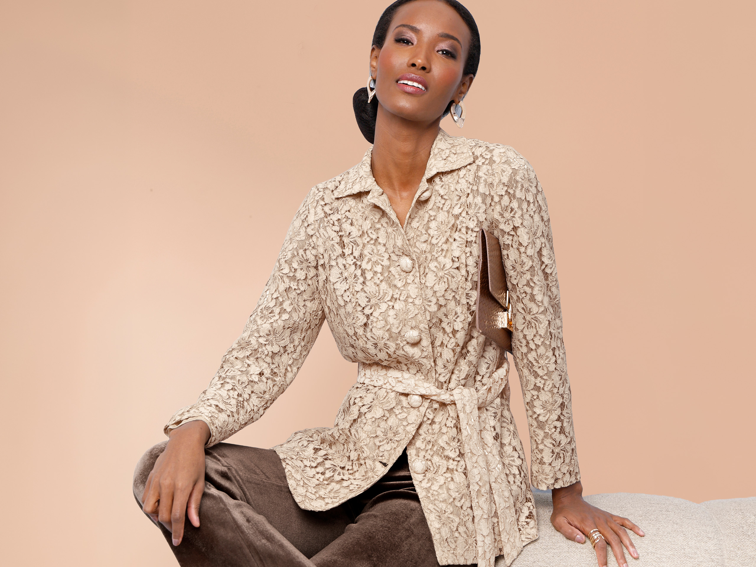 Ein Model trägt eine cremefarbene, strukturierte Jacke mit Gürtel und Knöpfen sowie eine braune Hose. Der Hintergrund ist beige.