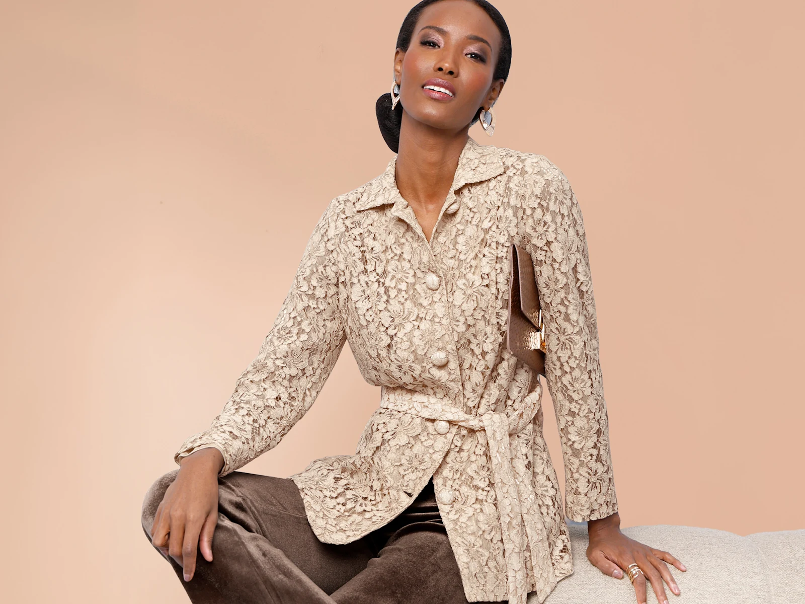 Ein Model trägt eine cremefarbene, strukturierte Jacke mit Gürtel und Knöpfen sowie eine braune Hose. Der Hintergrund ist beige.