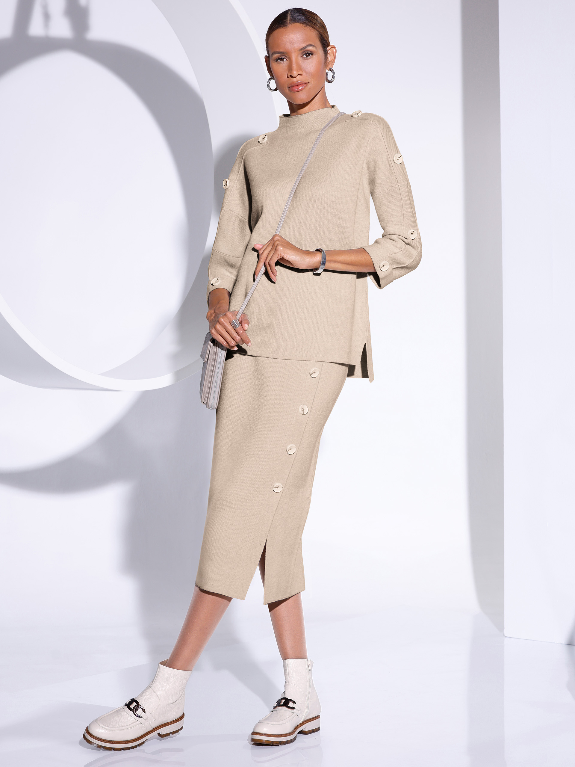 Ein Model trägt ein beiges, strukturiertes Oberteil mit drei Viertelärmeln und Knöpfen an den Ärmeln, kombiniert mit einem passenden, knielangen Rock. Die weißen Stiefel haben eine robuste Sohle. Der Hintergrund ist hell und minimalistisch gestaltet.