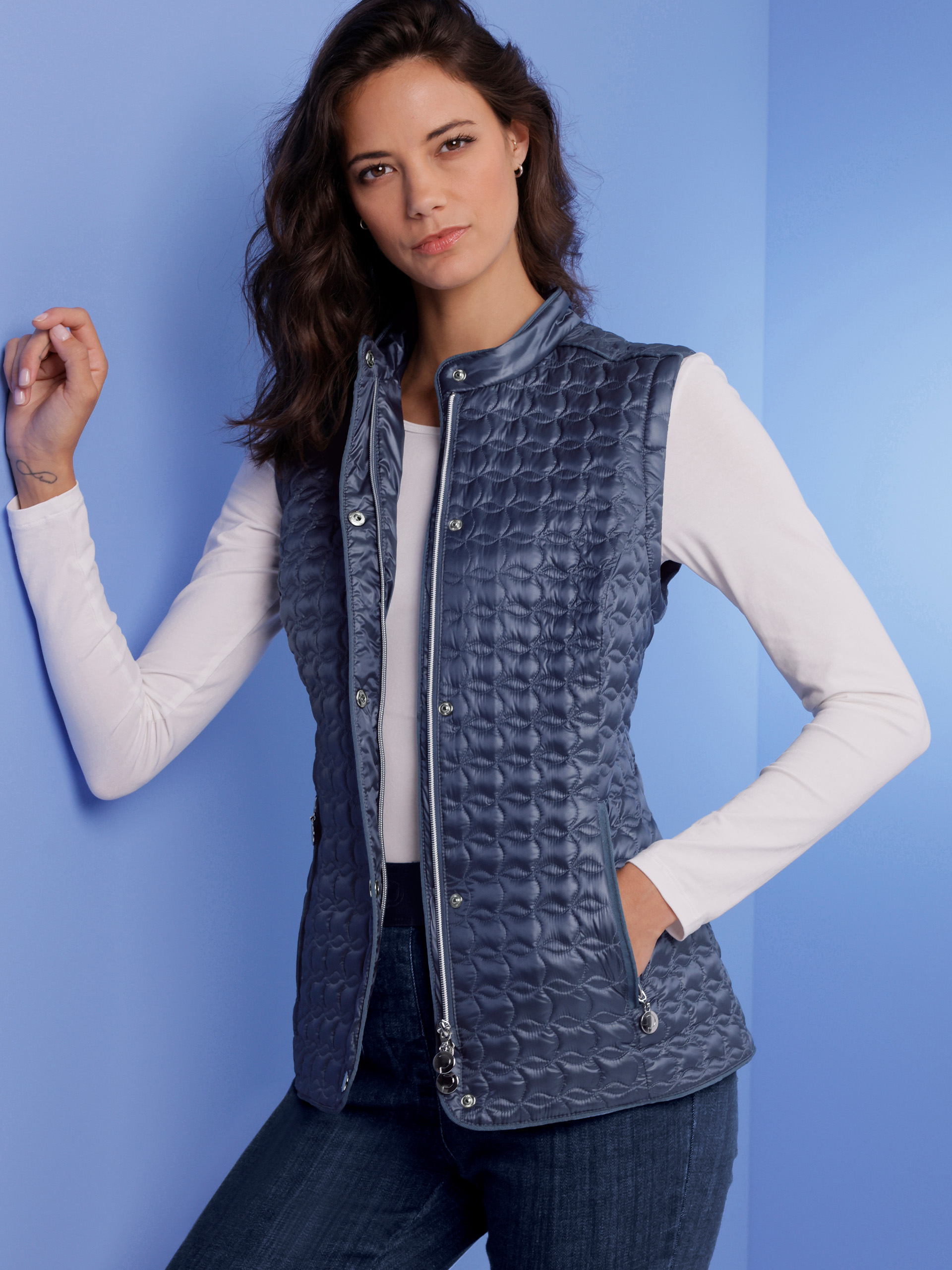 Ein Model lehnt an einer blauen Wand, trägt eine gesteppte, dunkelblaue Weste über einem weißen Langarmshirt und dunkle Jeans.