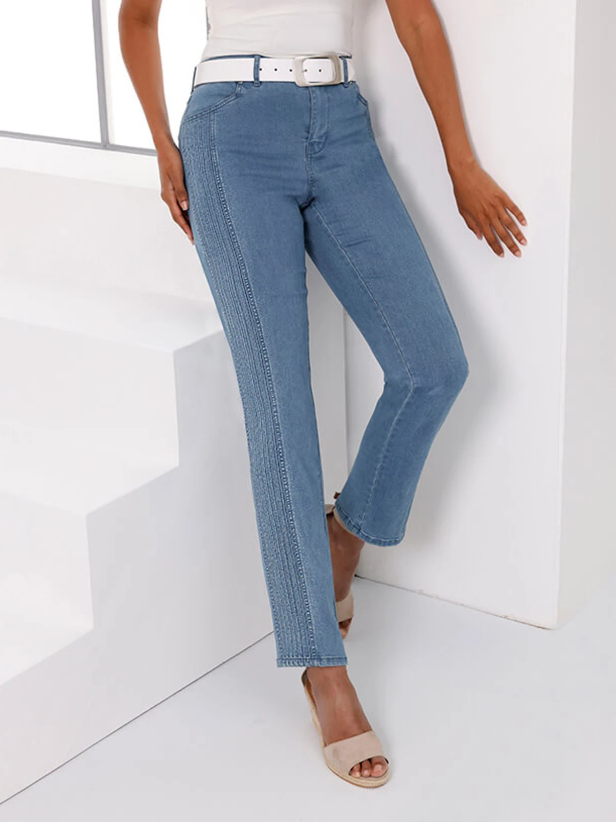 Ein Model trägt eine weiße Bluse und blaue Jeans, während es an einer Treppe steht.