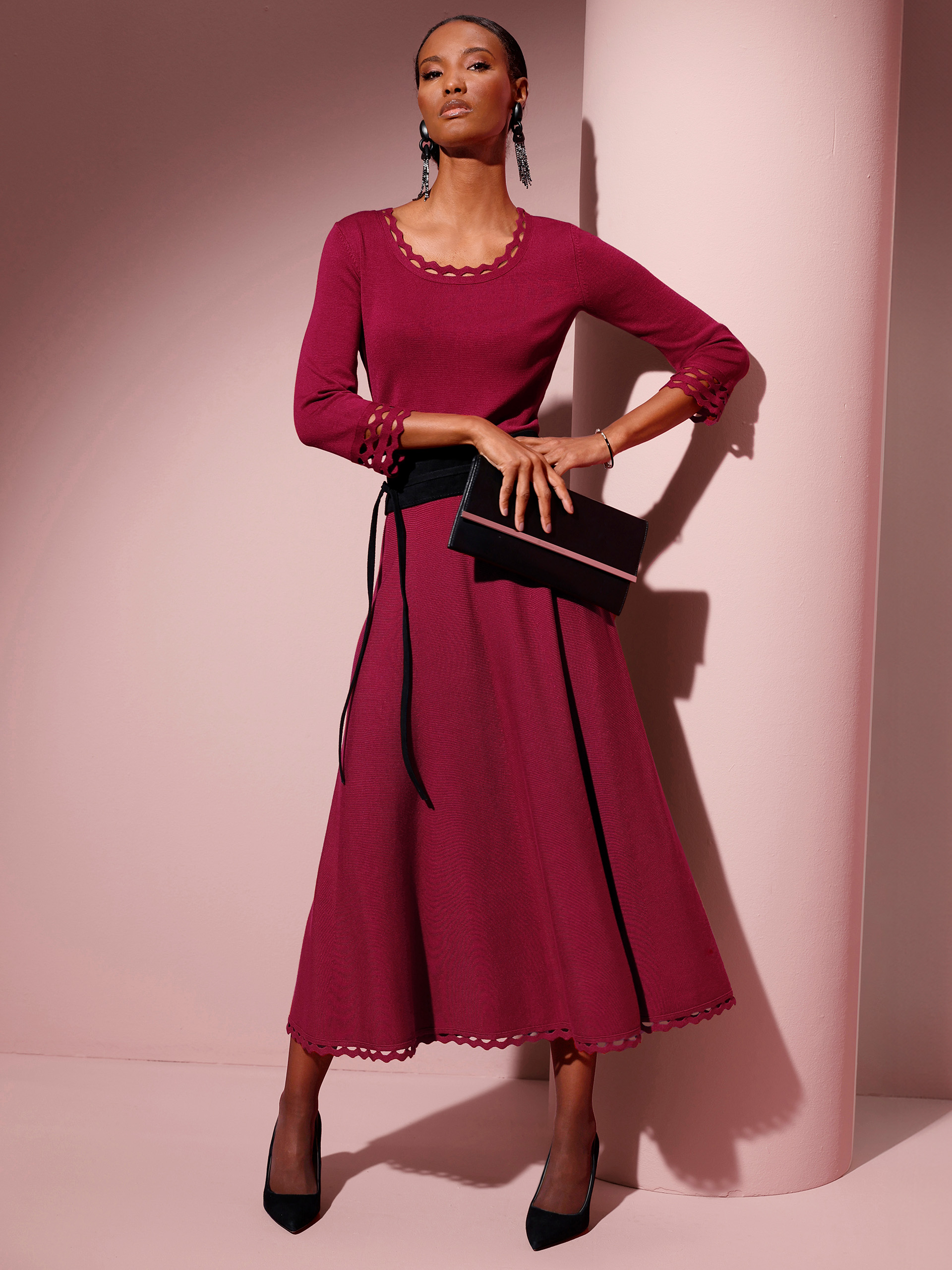 Ein Model trägt ein rotes, langärmliges Oberteil, dazu einen passenden Rock mit einem schwarzen Gürtel und einer schwarzen Clutch. Der Hintergrund ist rosa.
