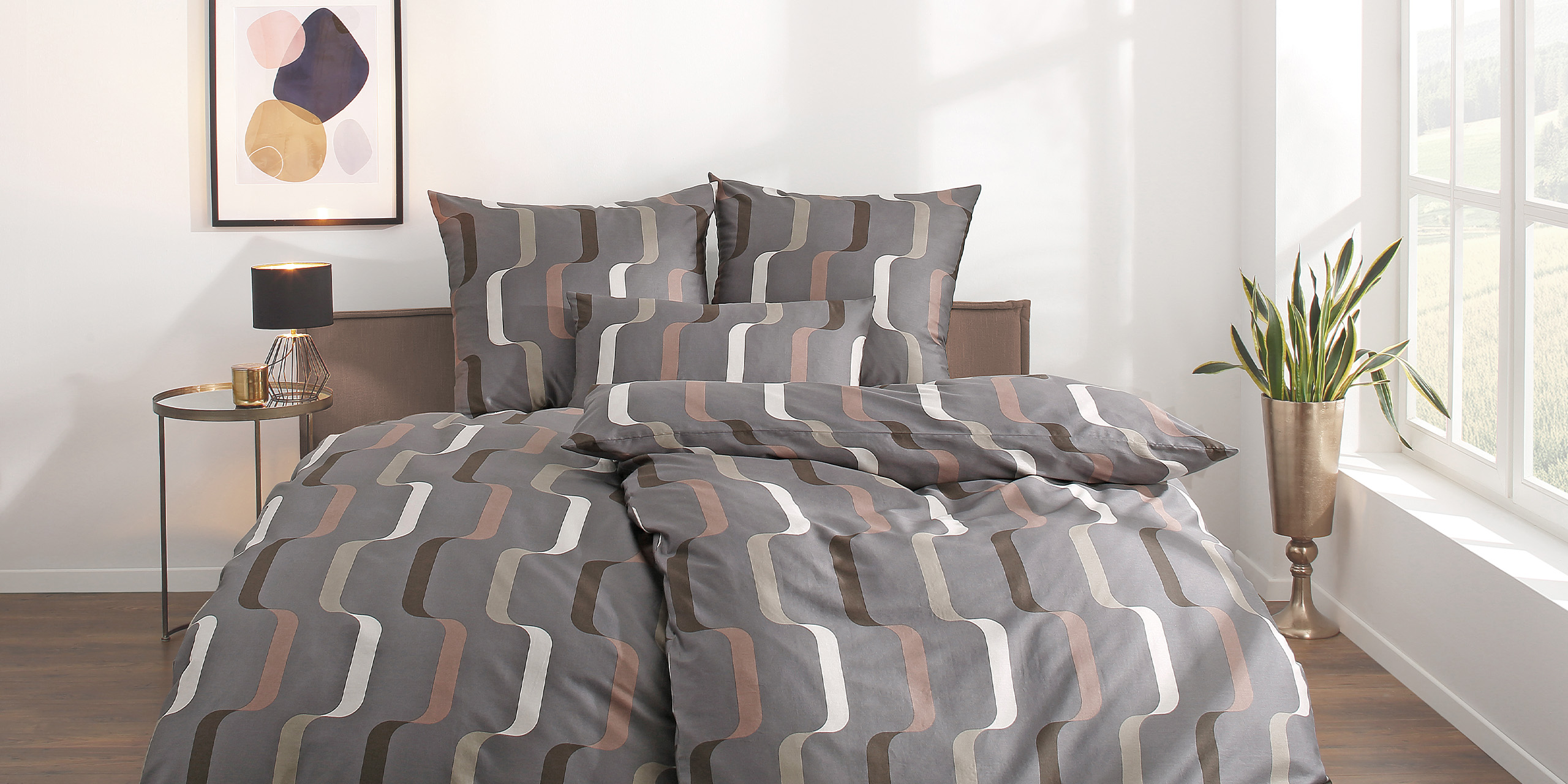 Bett mit gemusterter Bettwäsche in Grau und Braun, daneben Nachttisch und Pflanze.