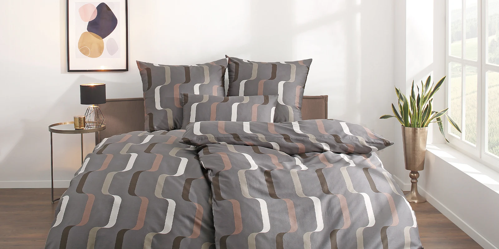 Bett mit gemusterter Bettwäsche in Grau und Braun, daneben Nachttisch und Pflanze.