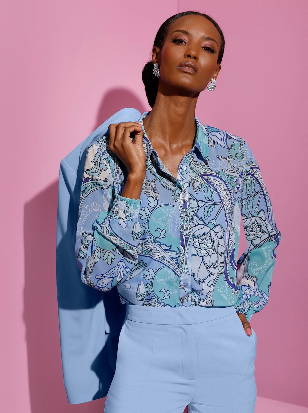 Model stehend vor einem pinken Hintergrund. Das Model trägt eine blaue Hose und eine blaue Bluse mit floralem Print.