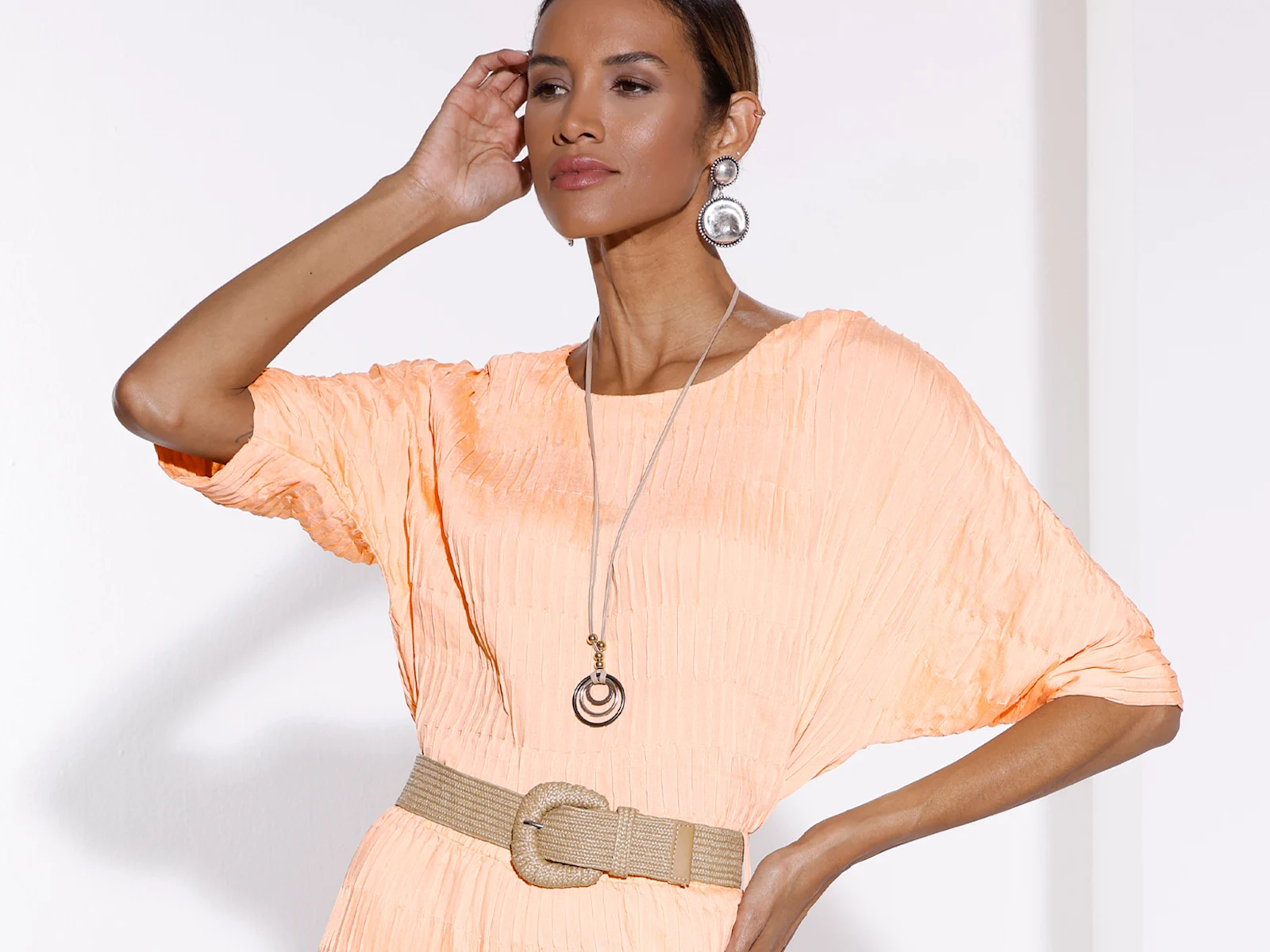 Ein Model trägt ein apricotfarbenes, strukturiertes Kleid mit breitem Gürtel, auffälligen Ohrringen und langer Kette vor weißem Hintergrund.