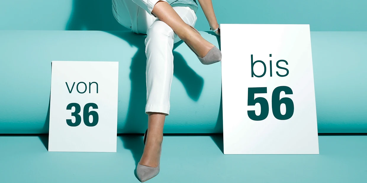 Frau in weißer Hose und grauen Schuhen sitzt vor türkisfarbenem Hintergrund, flankiert von Schildern mit den Größenangaben 'von 36' und 'bis 56'. Frau in weißer Hose und grauen Schuhen sitzt vor türkisfarbenem Hintergrund, flankiert von Schildern mit den Größenangaben 'von 36' und 'bis 56'.