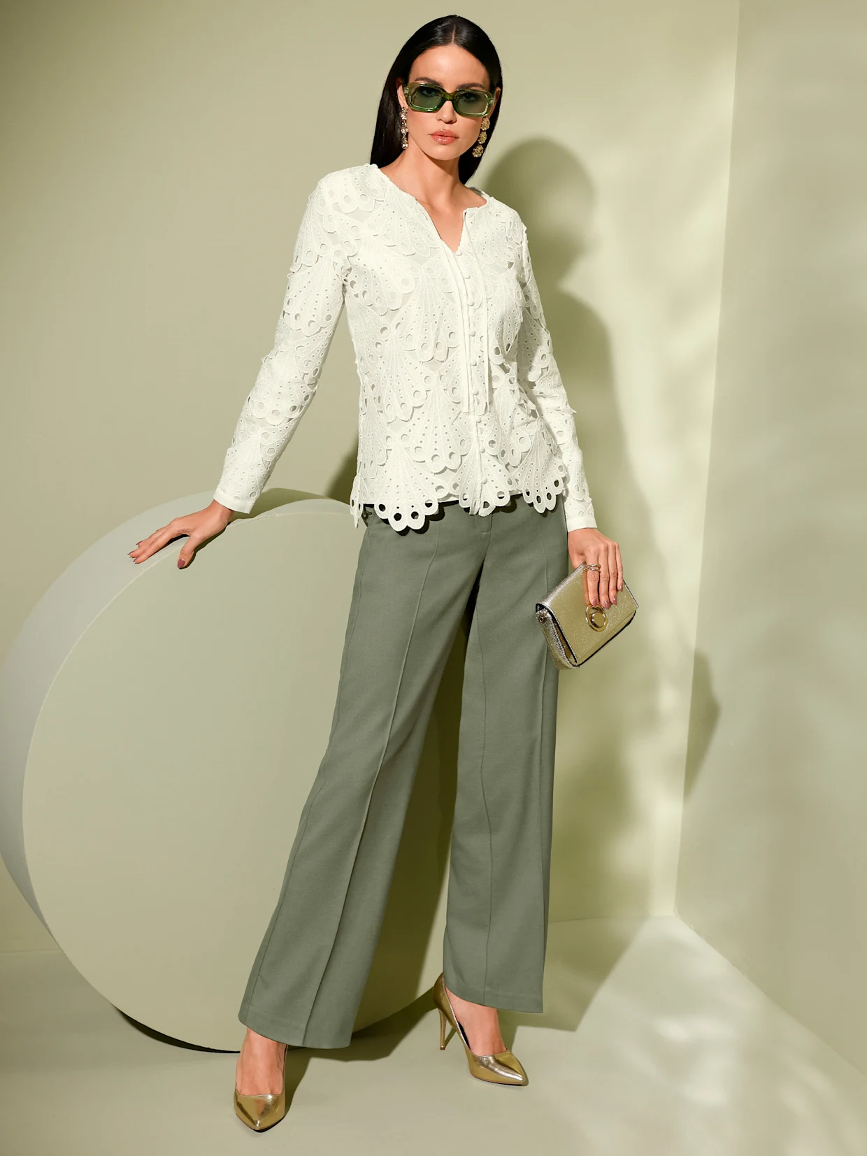 Ein Model trägt eine weiße, strukturierte Bluse und olivgrüne, weite Hosen. Sie hält eine goldene Clutch. Die Schuhe sind goldfarben. Der Hintergrund ist grün. Ein Model trägt eine weiße, strukturierte Bluse und olivgrüne, weite Hosen. Sie hält eine goldene Clutch. Die Schuhe sind goldfarben. Der Hintergrund ist grün.