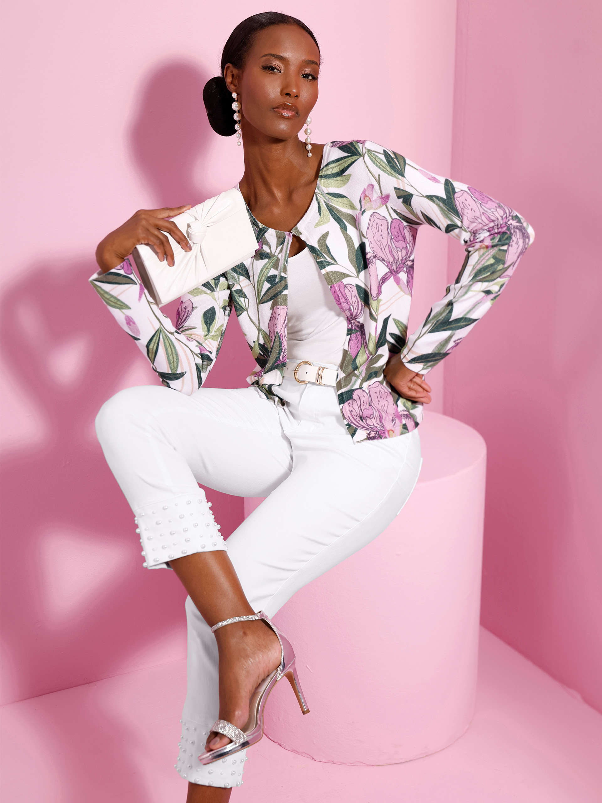 Ein Model sitzt vor rosa Hintergrund, trägt weiße Hose mit Perlenbesatz, florale Jacke in Rosa-Grün, silberne High Heels und hält eine Clutch.