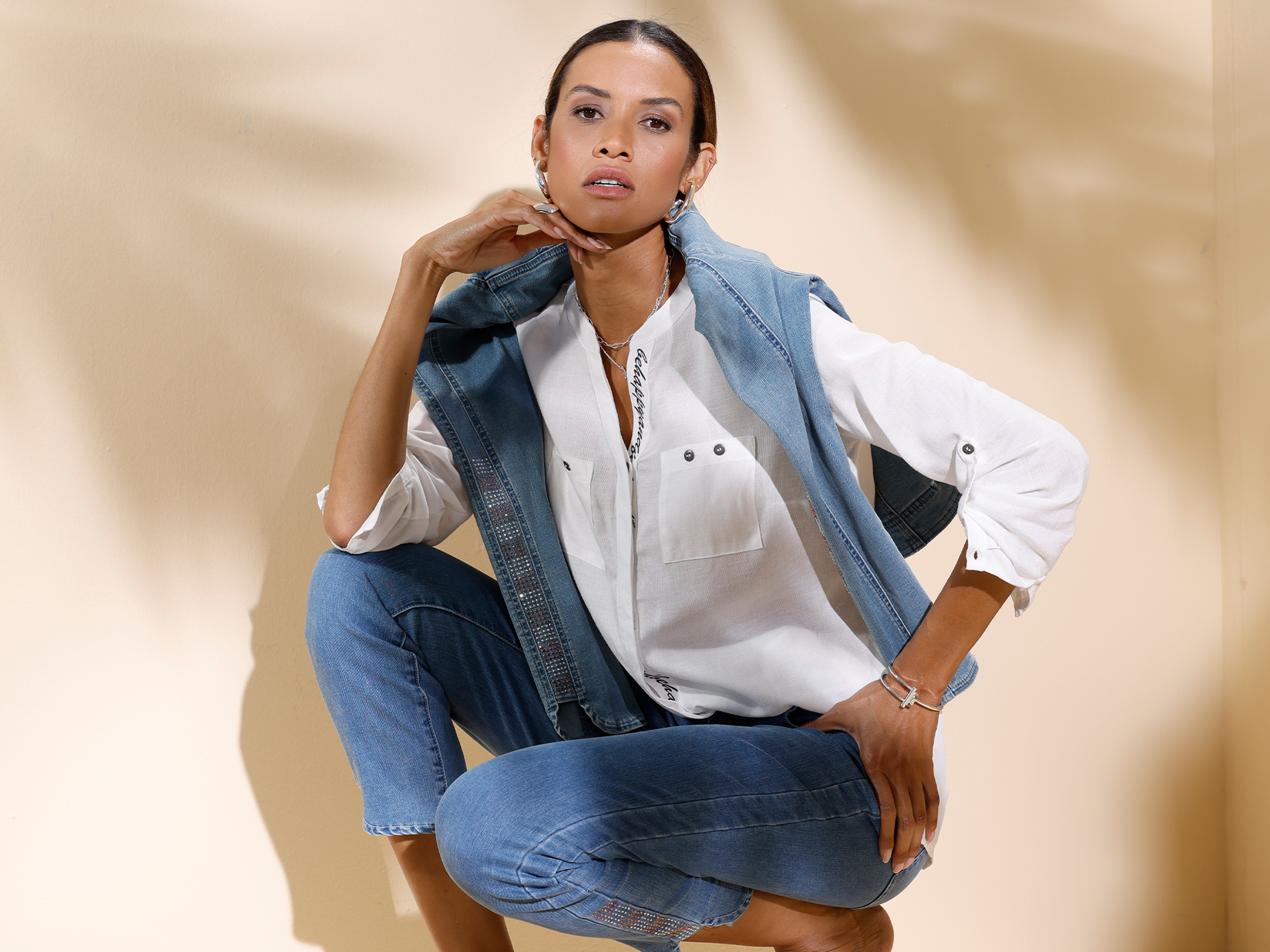 Ein Model sitzt lässig vor hellem Hintergrund, trägt eine weiße Bluse, blaue Jeans und eine aufgelegte Jeansjacke