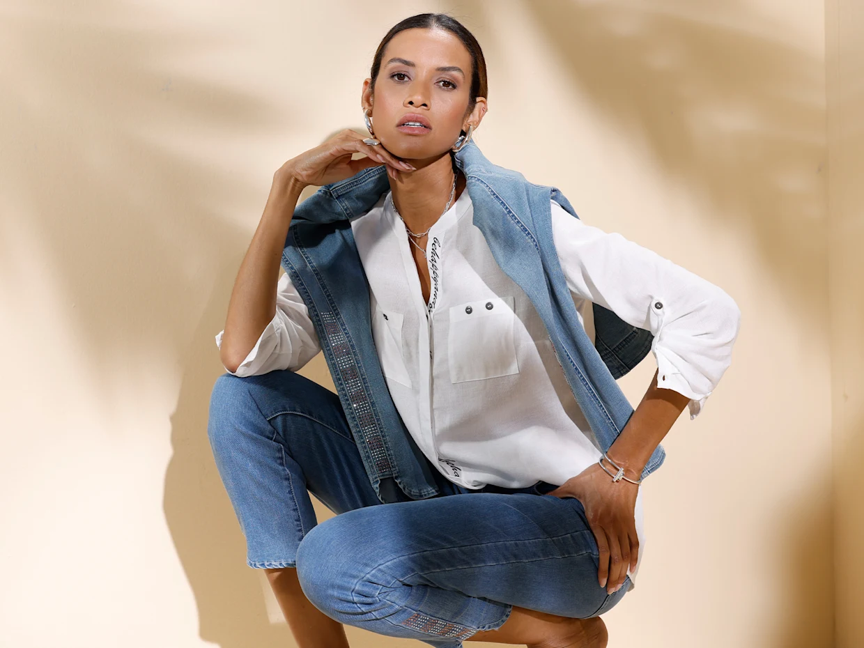 Ein Model sitzt lässig vor hellem Hintergrund, trägt eine weiße Bluse, blaue Jeans und eine aufgelegte Jeansjacke