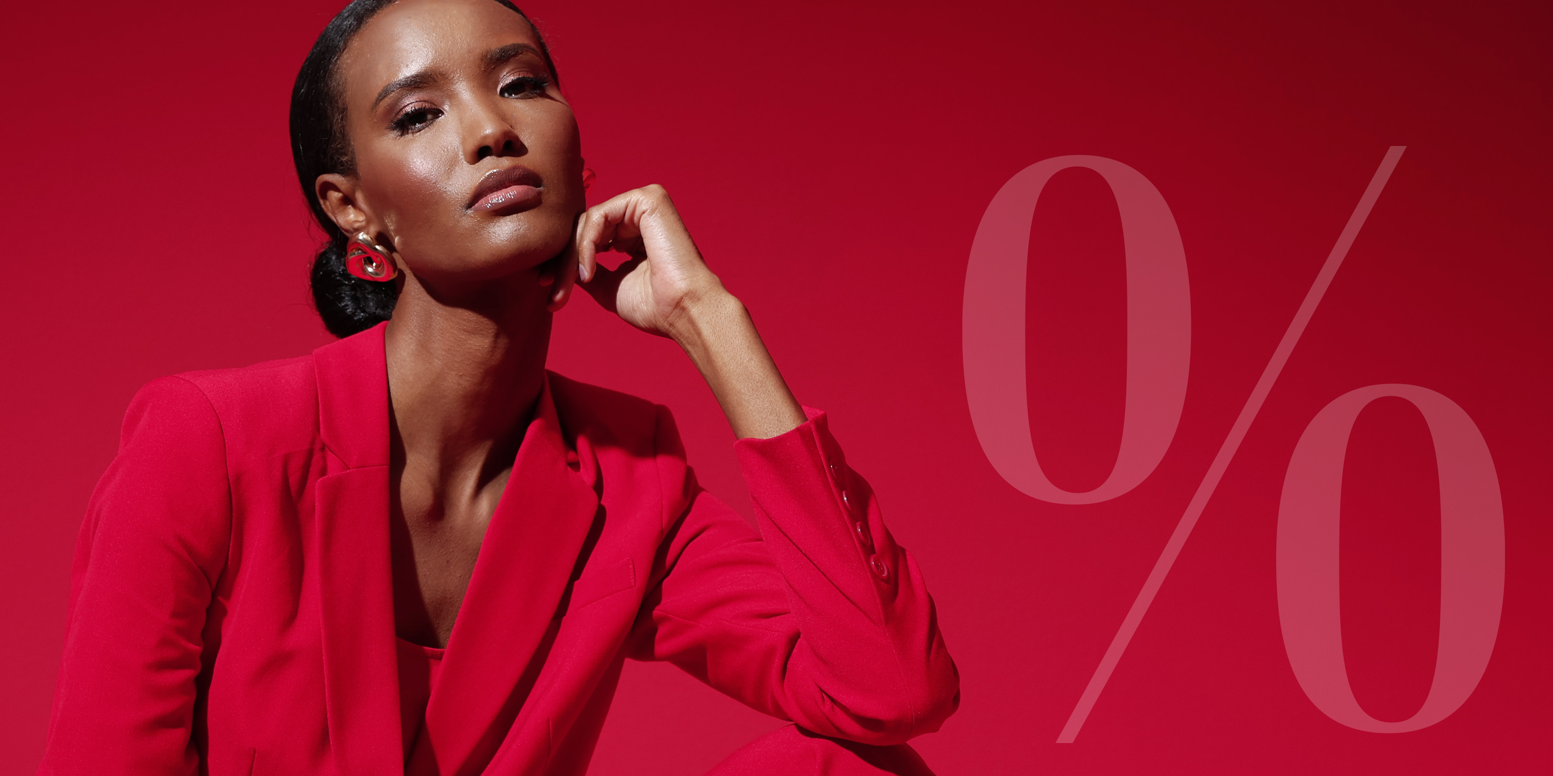 Ein Model sitzt vor rotem Hintergrund in einem roten Anzug, rechts im Bild ein großes, transparentes Prozentzeichen.