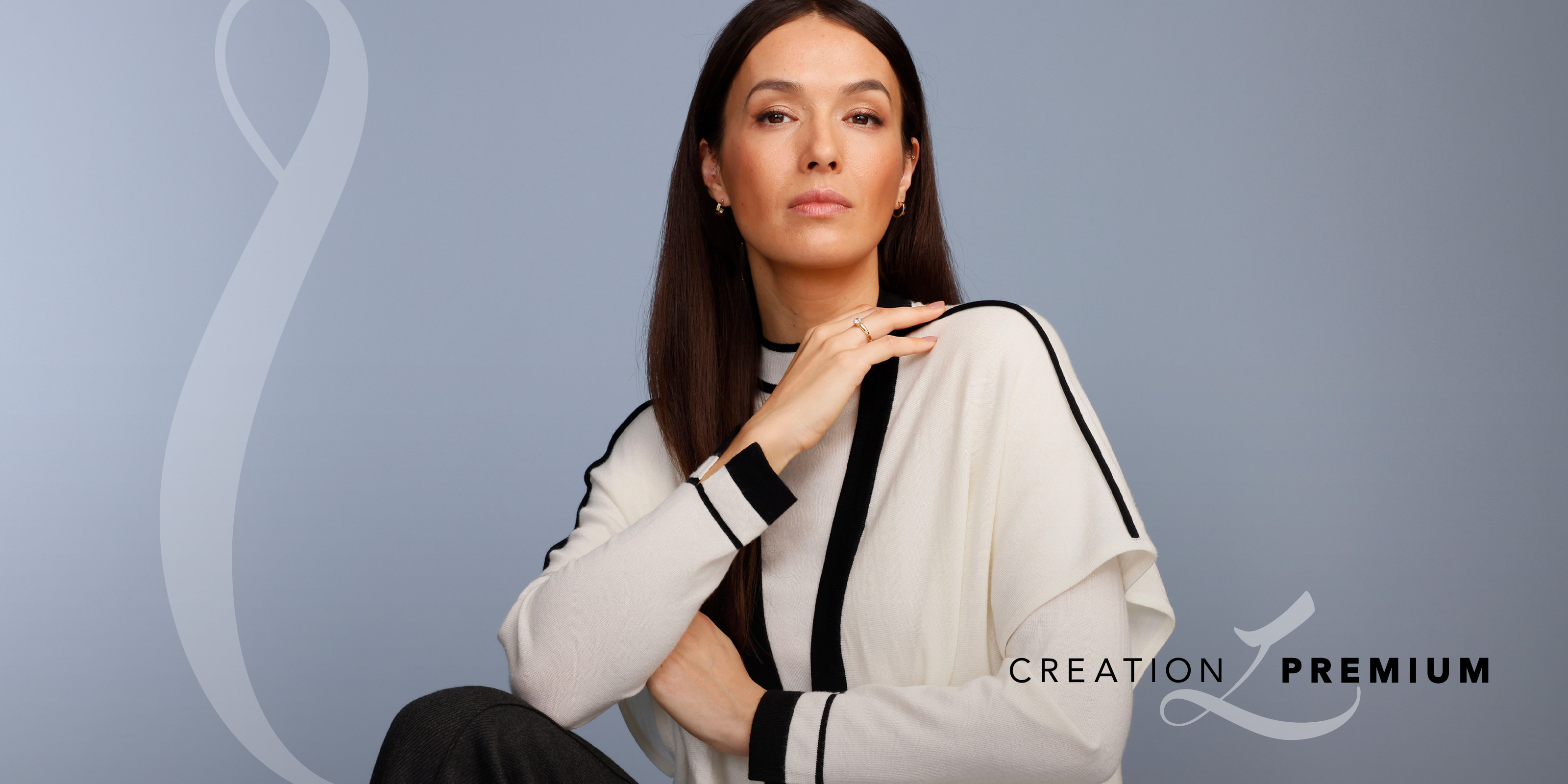 Ein Model trägt einen cremefarbenen Pullover mit schwarzen Streifen, sitzt vor graublauem Hintergrund. Text: 'Creation Premium'.