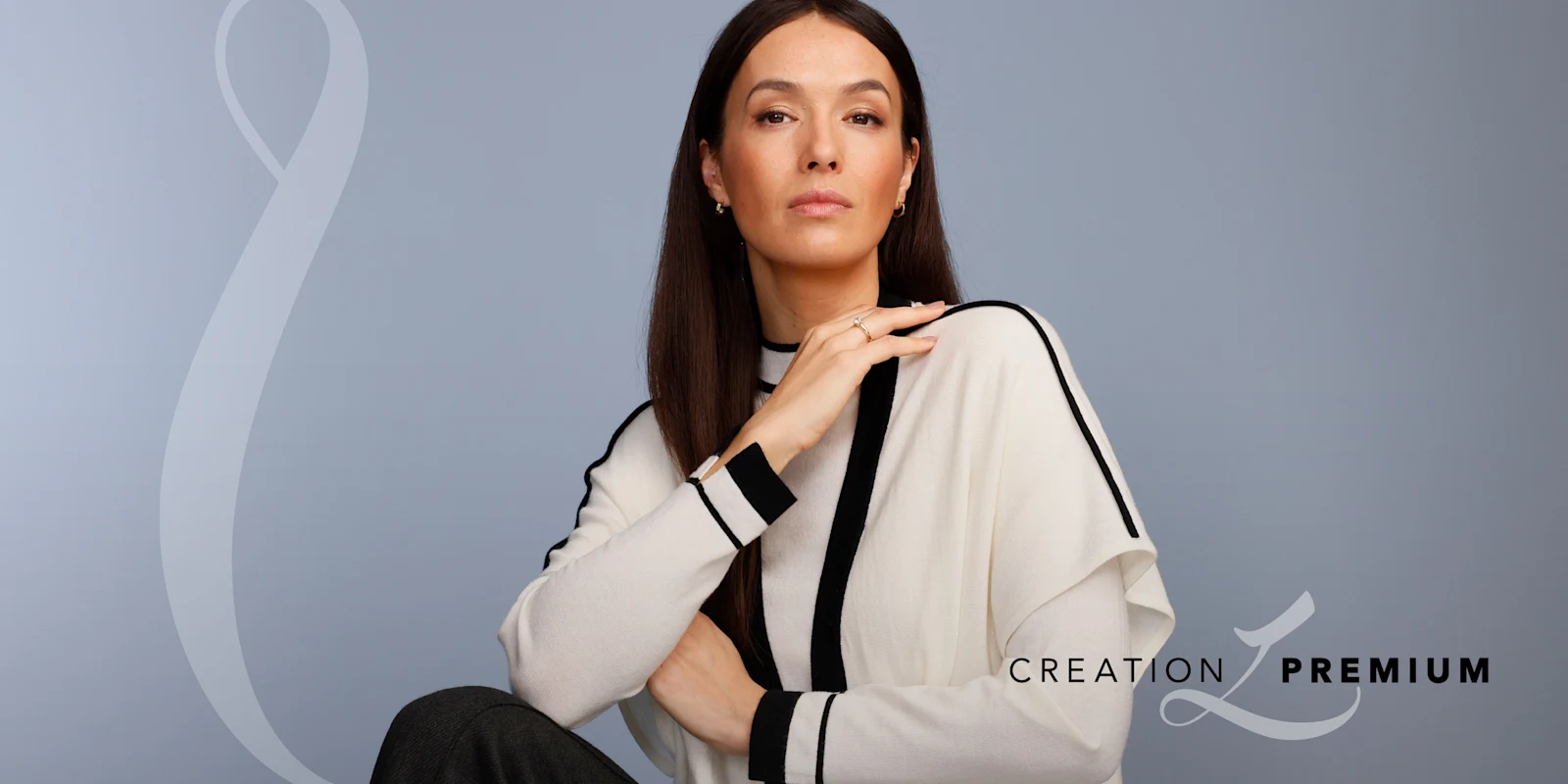 Ein Model trägt einen cremefarbenen Pullover mit schwarzen Streifen, sitzt vor graublauem Hintergrund. Text: 'Creation Premium'.
