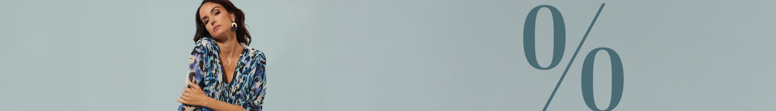 Ein Model sitzt auf einem Würfel, trägt eine gemusterte Tunika in Blau- und Beigetönen sowie dunkle Hose. Großes Prozentzeichen im Hintergrund.