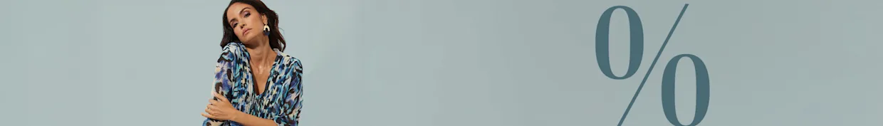 Ein Model sitzt auf einem Würfel, trägt eine gemusterte Tunika in Blau- und Beigetönen sowie dunkle Hose. Großes Prozentzeichen im Hintergrund.